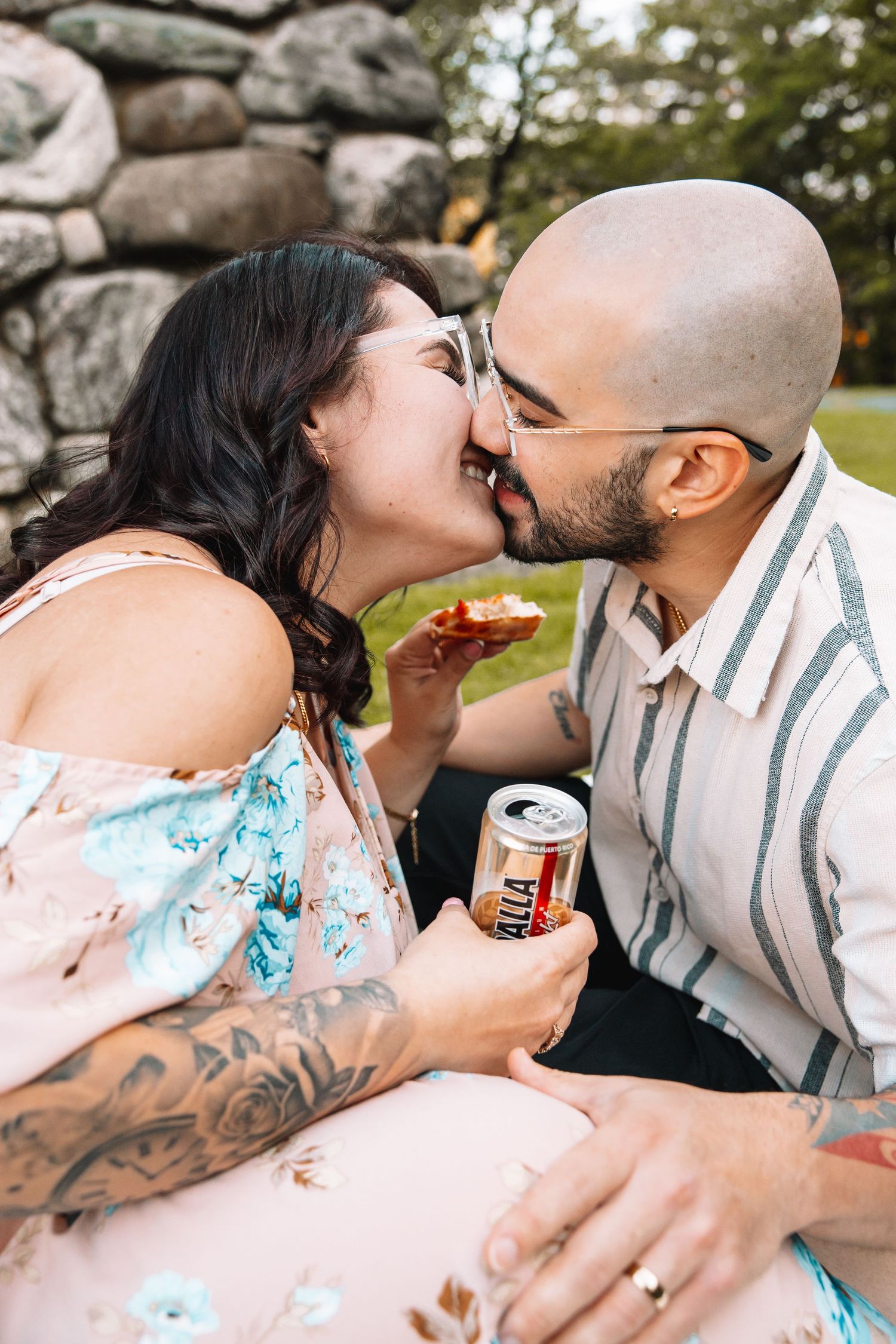Couple sharing a romantic kiss during a picnic, holding a beer can and pizza slice outdoors.
