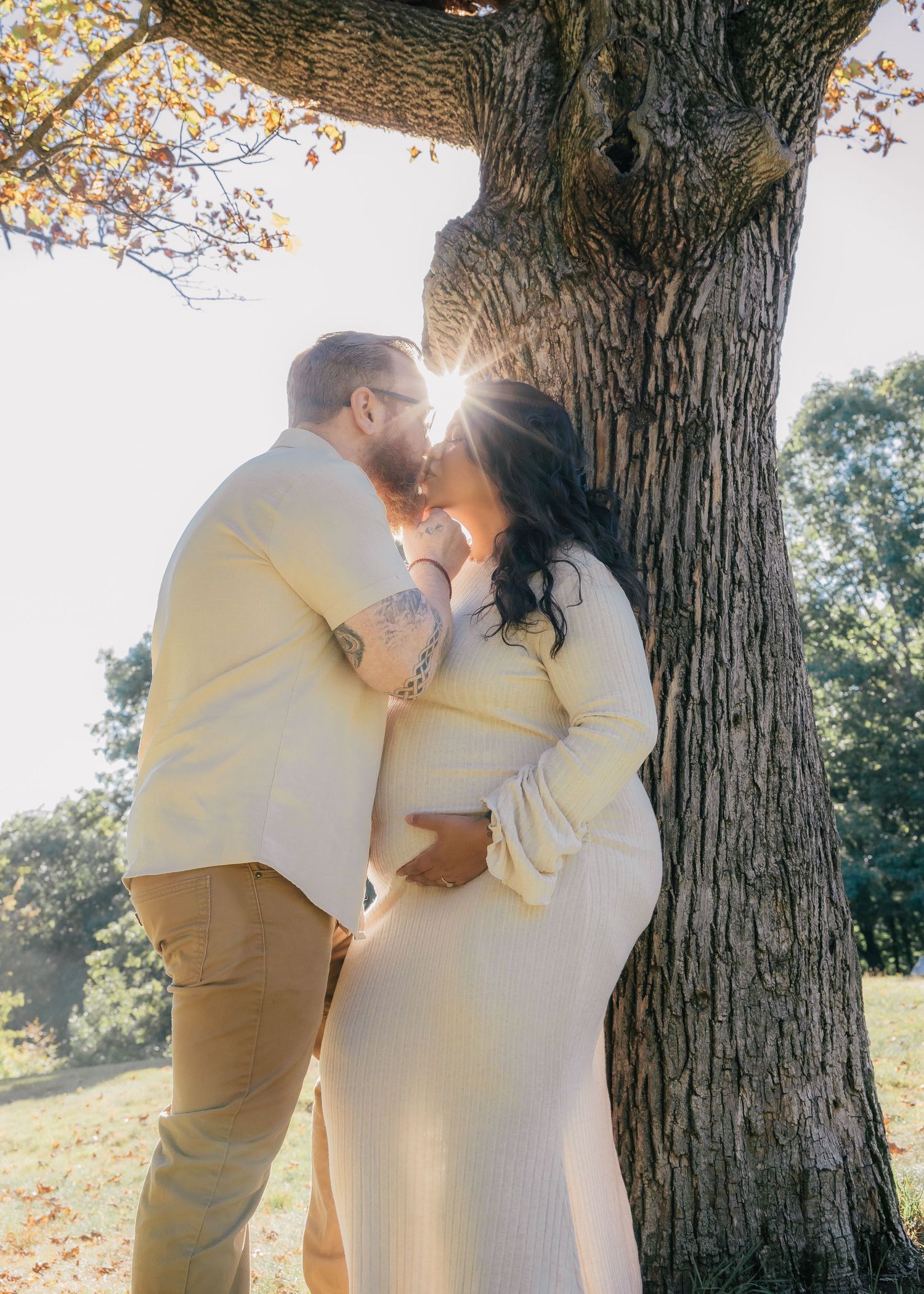 Couple sharing a kiss under a sunlit tree during a maternity photoshoot outdoors in autumn.