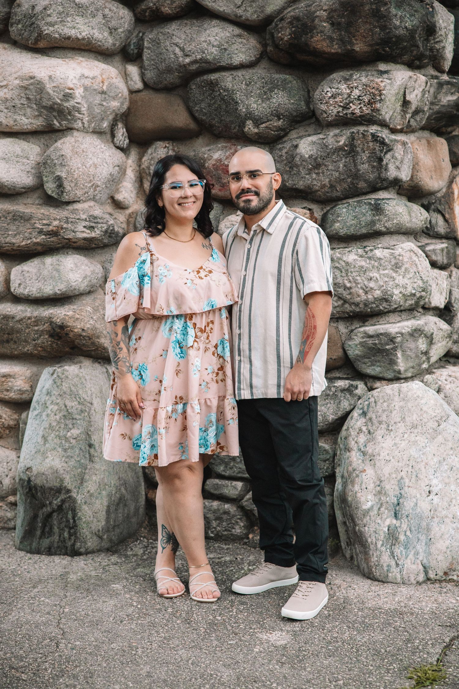 A couple poses smiling in front of a stone wall, woman in floral dress, man in striped shirt and dark pants.