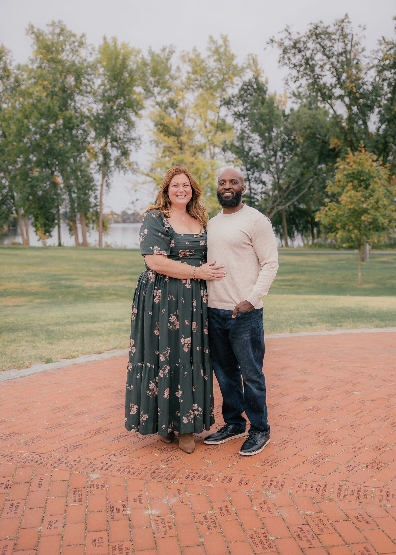 Couple posing together on a brick plaza in a park, woman in floral dress and man in beige sweater.