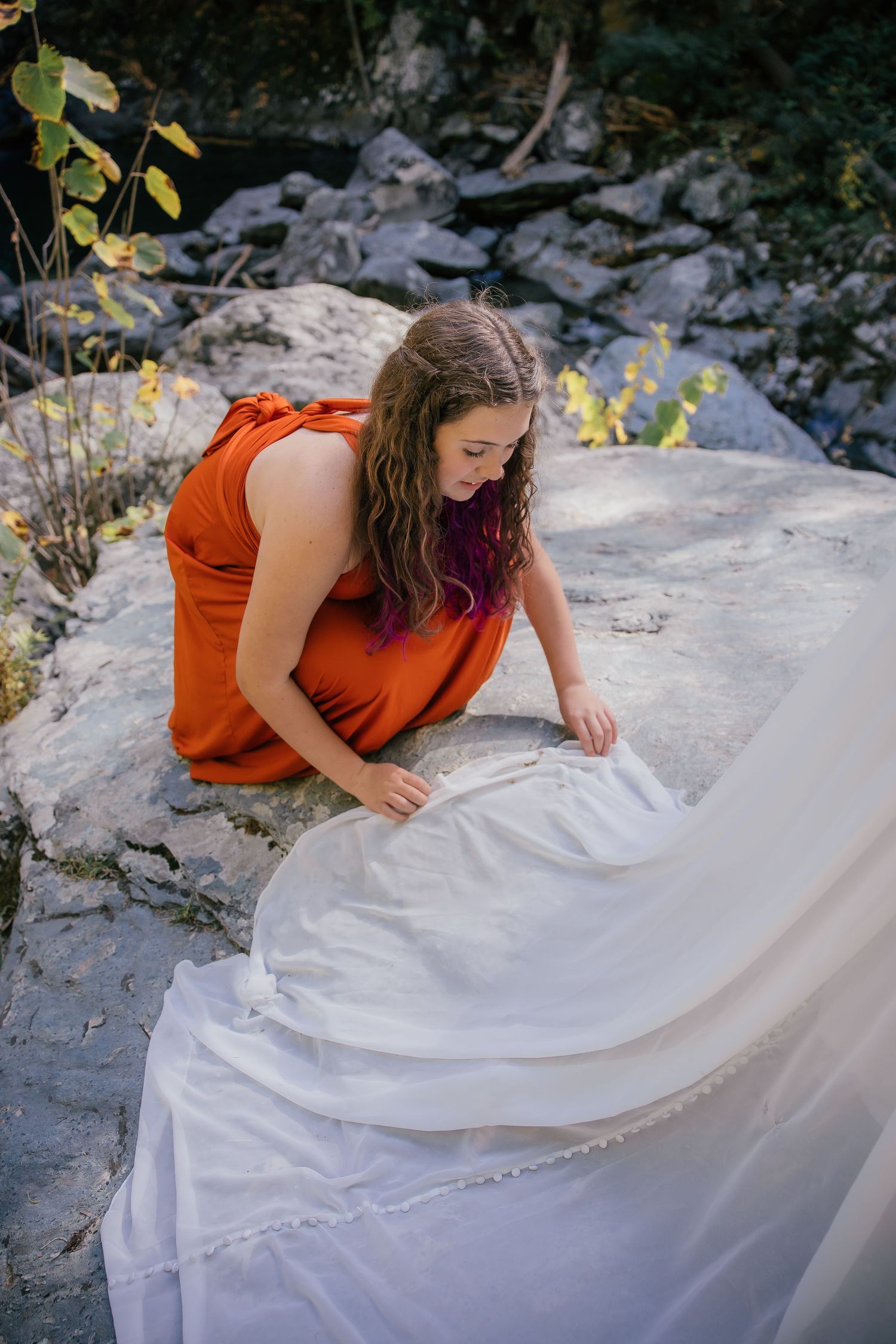Woman in orange dress kneeling on rocks adjusting a white wedding gown outdoors.