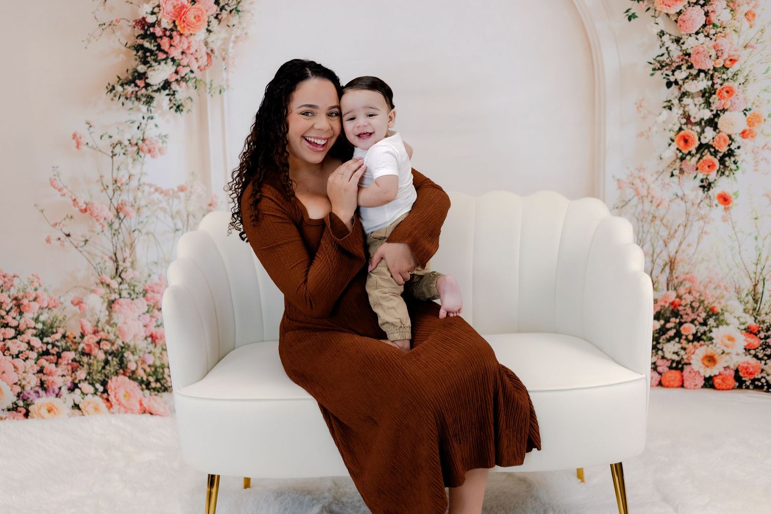 Happy woman in brown dress holding smiling baby on white sofa with floral backdrop.