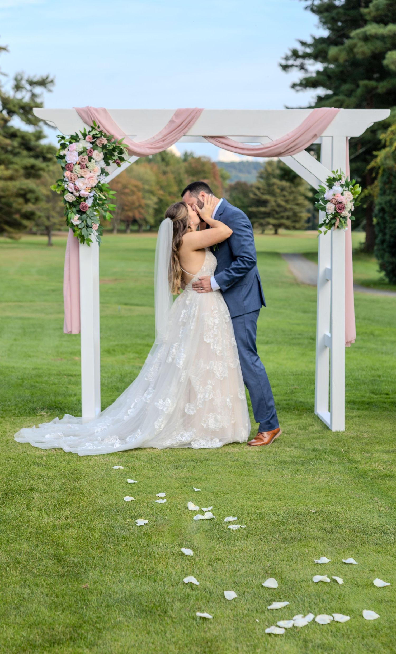 Newlyweds kiss under white arch decorated with pink fabric and flowers.