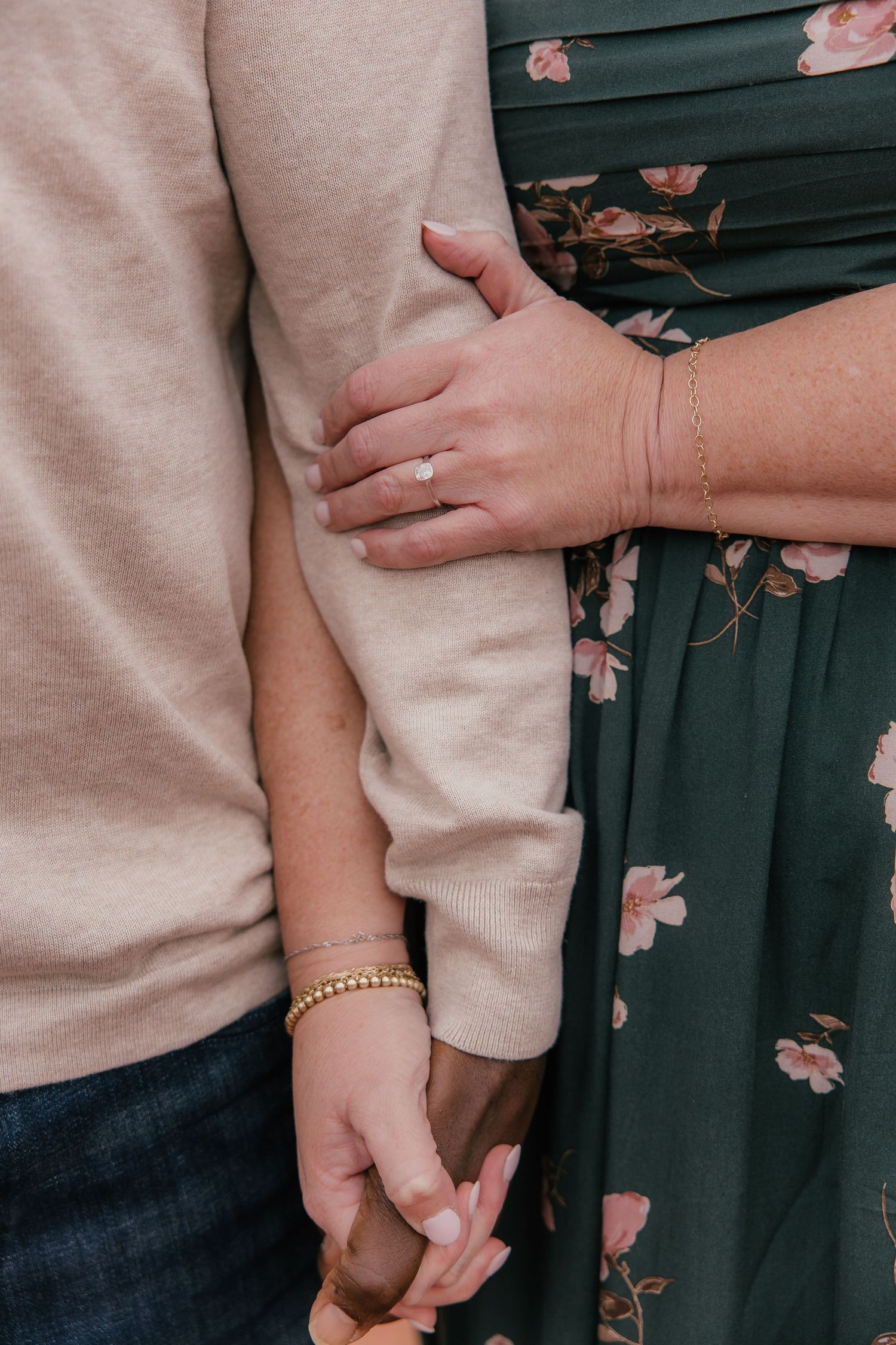 Close-up of couple holding hands, woman wearing engagement ring, gold bracelets, green floral dress, man in beige sweater.