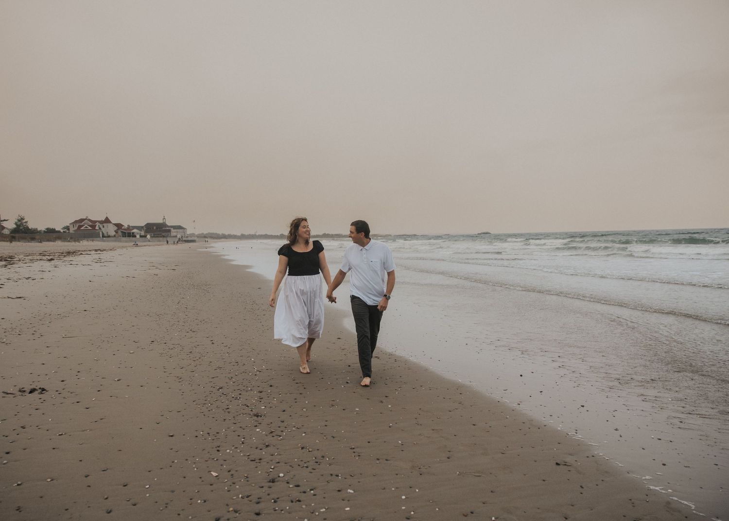 Couple holding hands and walking barefoot along a quiet sandy beach at dusk with gentle waves and a hazy sky.