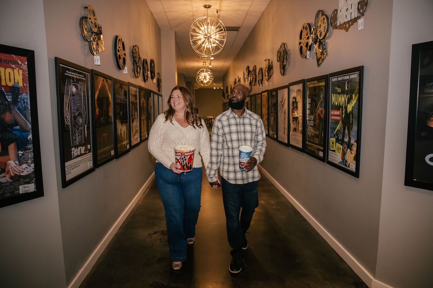 Two people hold drinks and walk side by side down a hallway lined with framed movie posters and decorative film reels on the walls.
