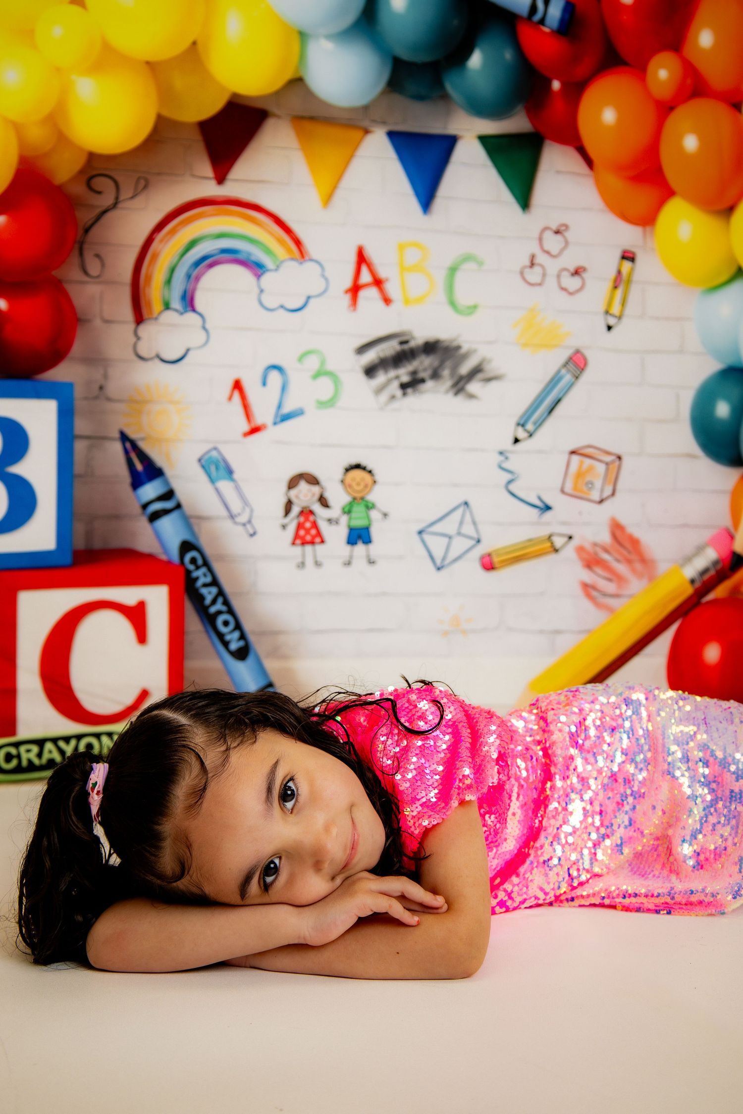 Young girl in pink sequin outfit poses on floor with colorful ABC school-themed balloon backdrop.