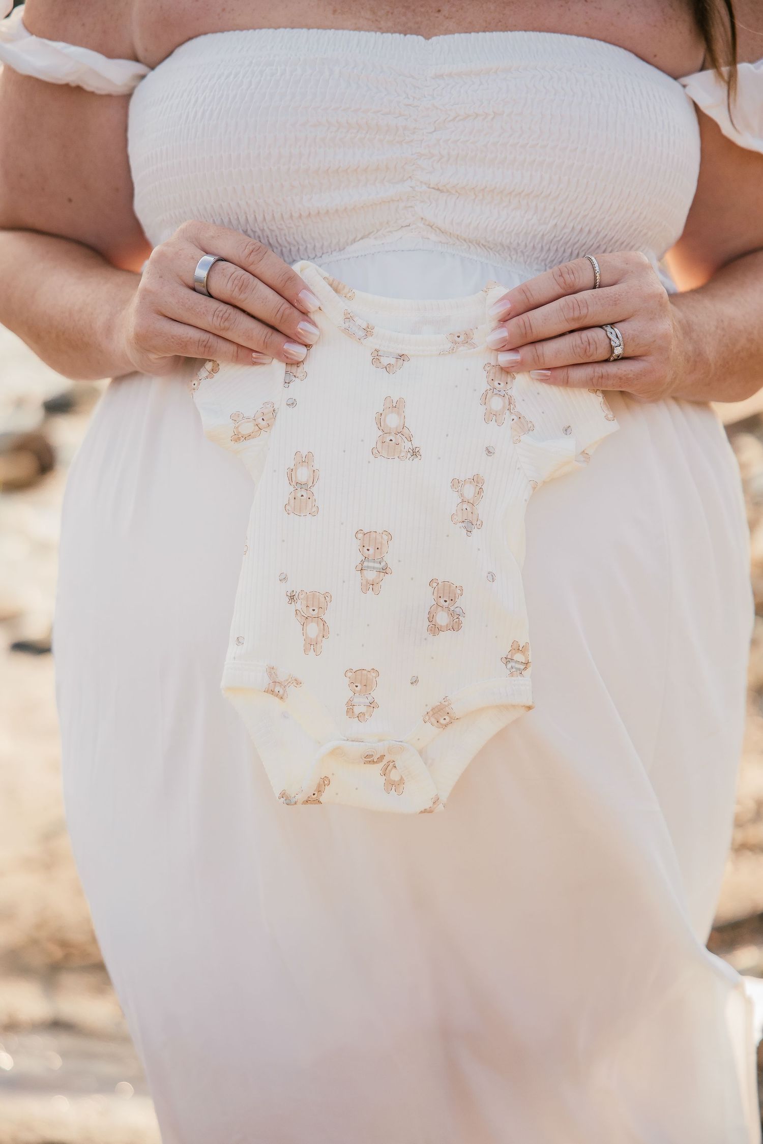Pregnant woman in white dress holding a cute bear-print baby onesie outdoors during a maternity photoshoot.