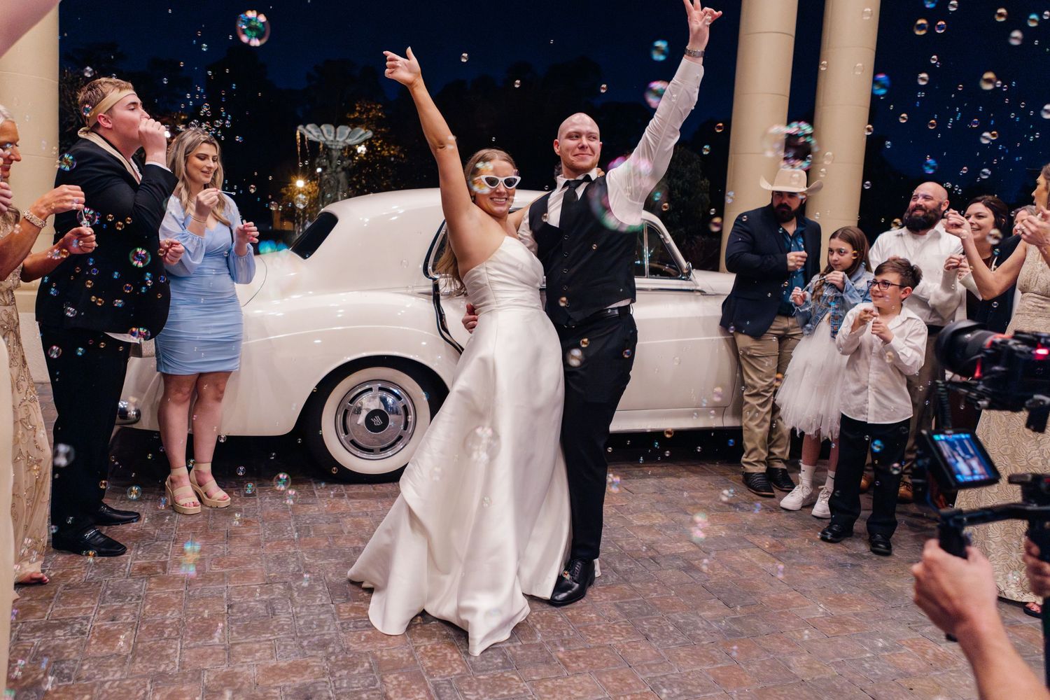Wedding celebration outside with vintage white car and floating bubbles at dusk.