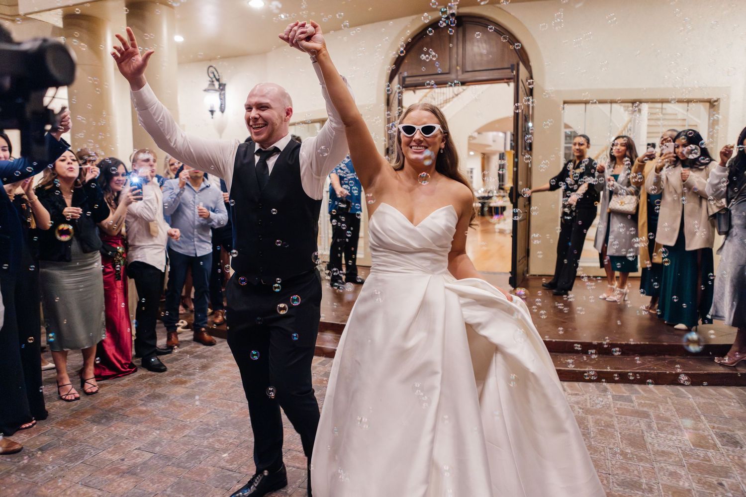 Newlyweds celebrate joyfully in elegant reception hall with guests and bubbles.