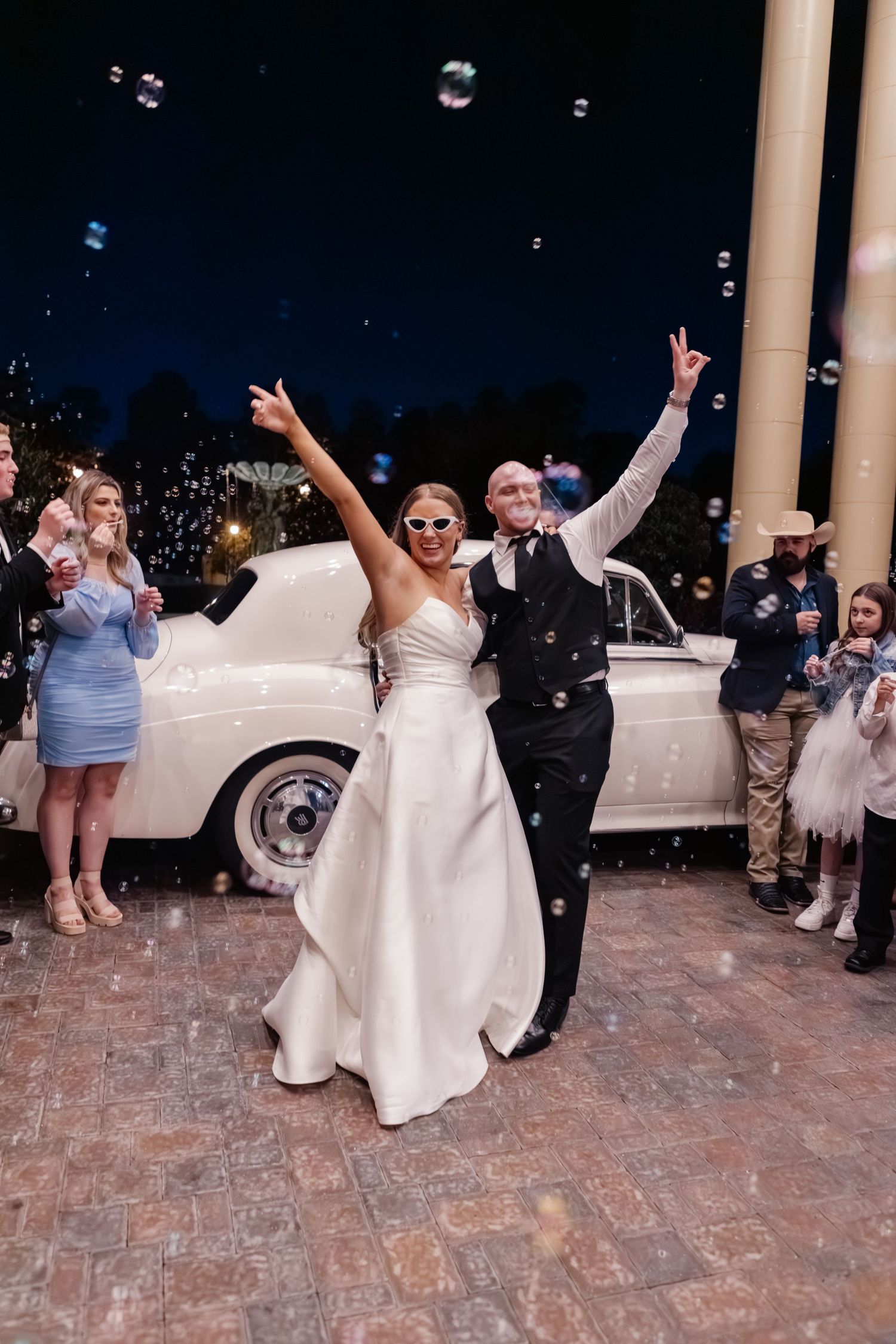Couple celebrates by vintage white car during evening wedding exit with bubbles.