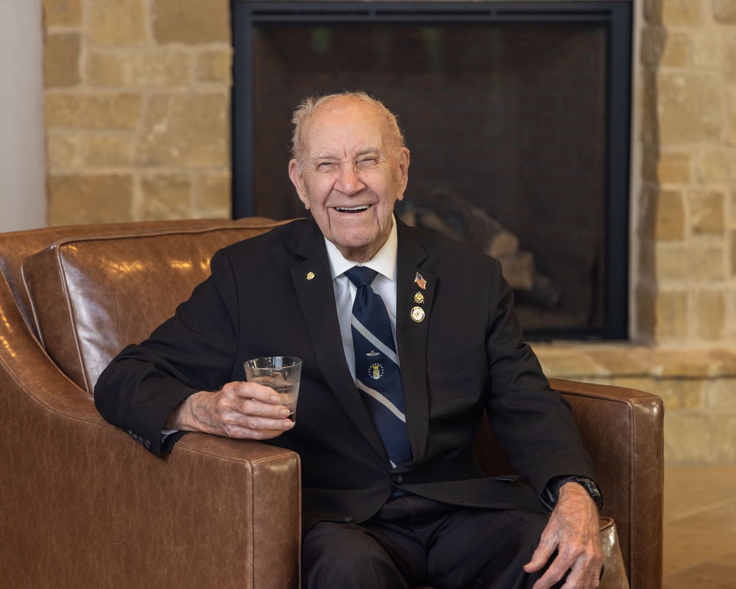 Elderly gentleman sits smiling in leather chair beside stone fireplace.