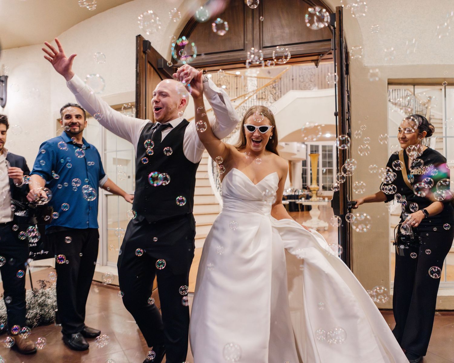 Wedding couple celebrates with guests while soap bubbles float through the air during indoor reception.