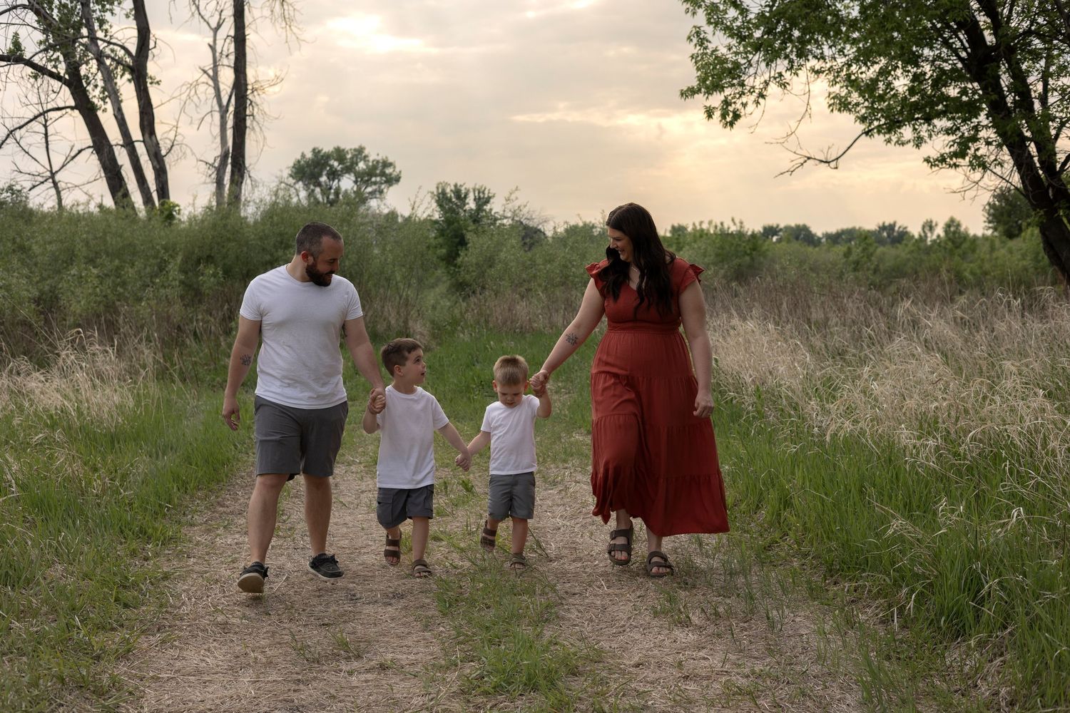 A family of four walks hand-in-hand along a dirt path through a scenic grassy landscape at dusk.