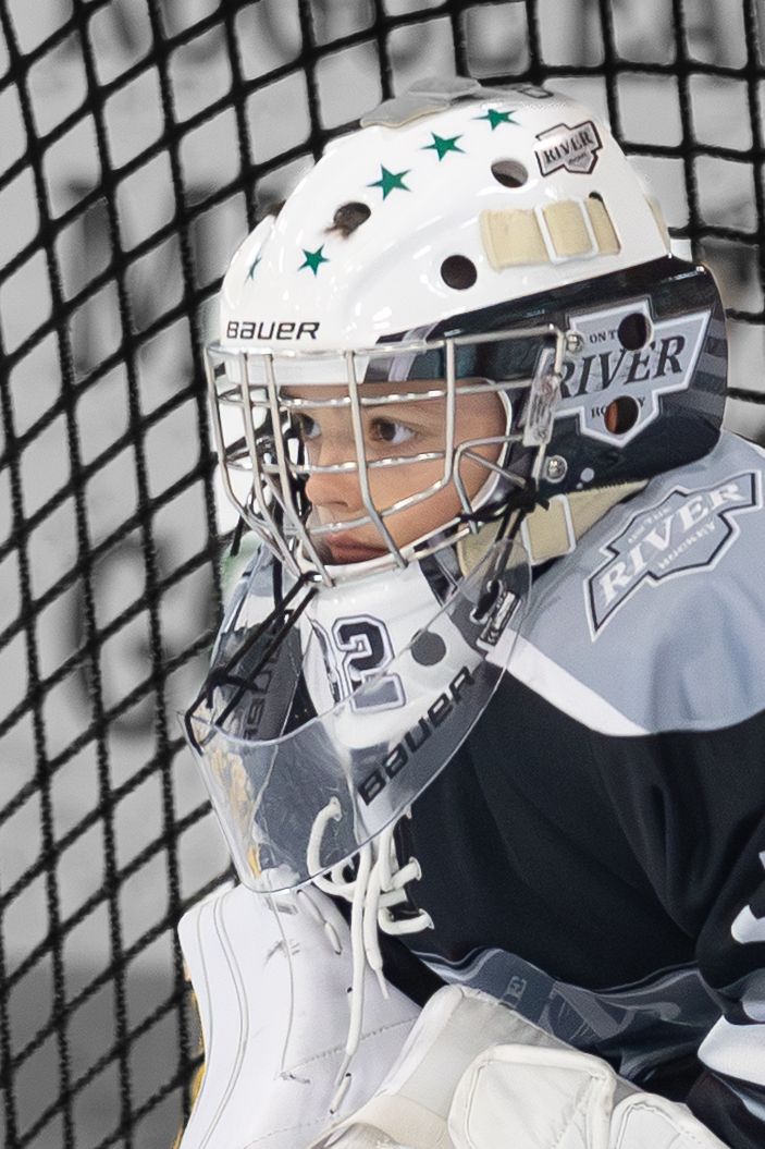 Young hockey goalie in Bauer helmet and River jersey positioned in front of net.