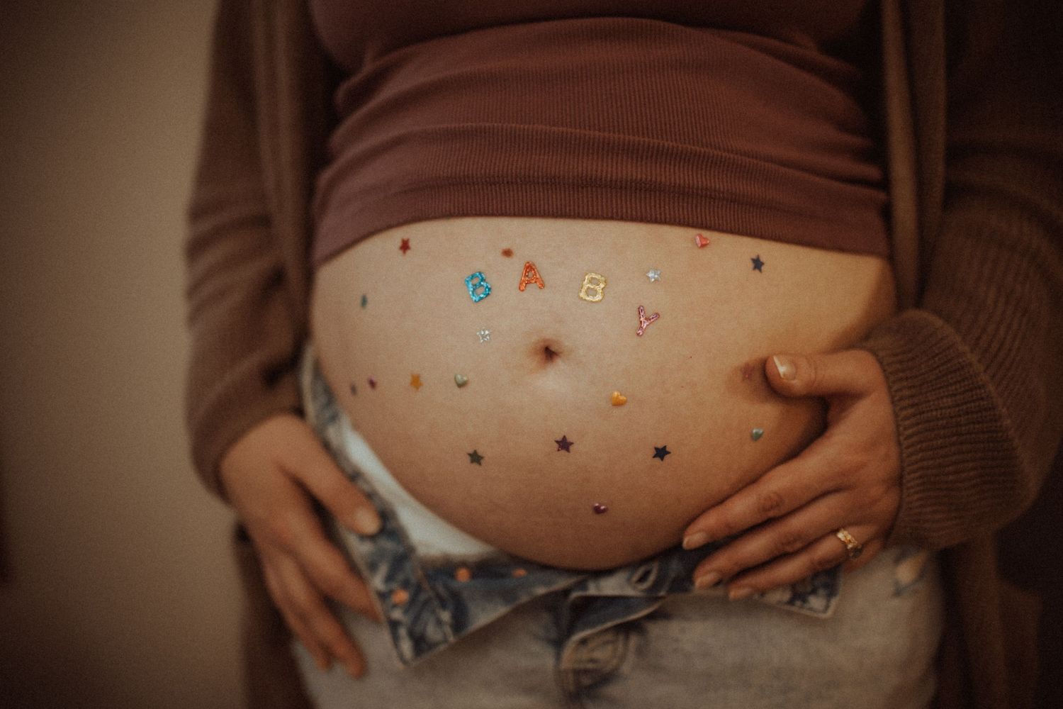 Close-up of a pregnant belly with colorful baby letter stickers and a partner's hand.