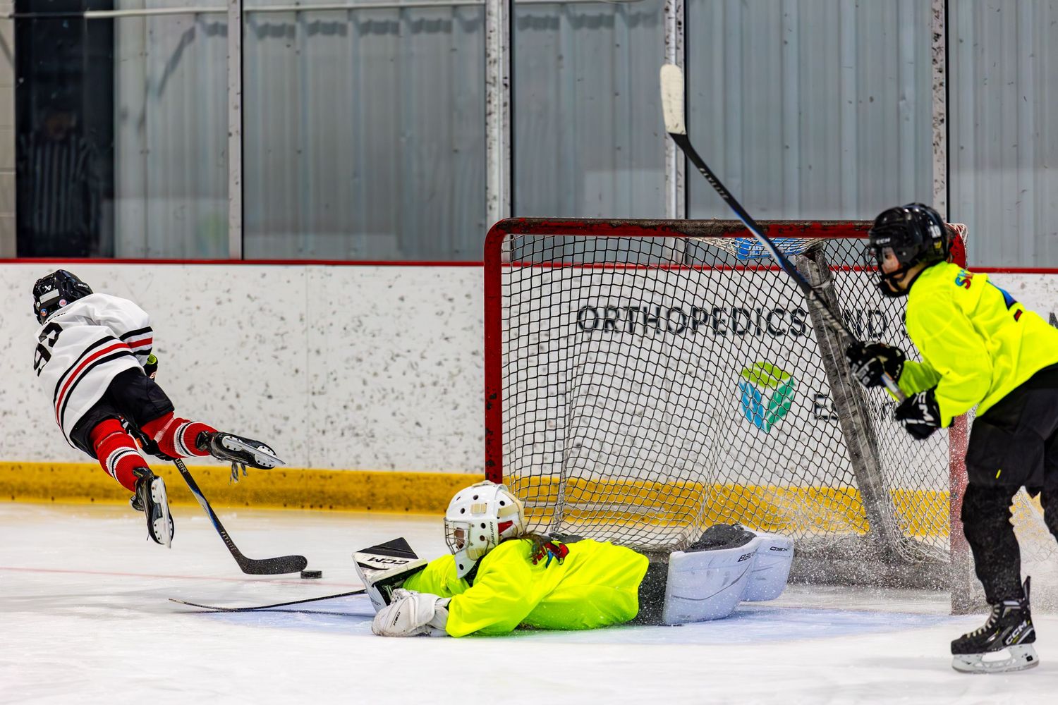 Youth ice hockey players in action near goal as goalie dives to make a save.