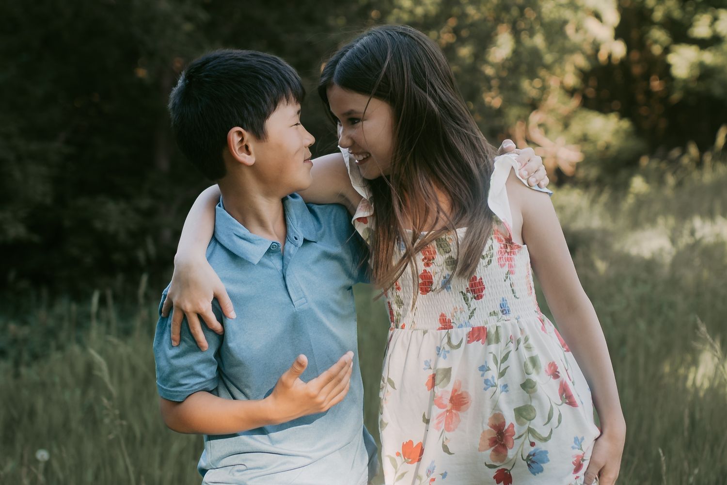 Two children hugging and smiling at each other in a sunlit meadow, boy in blue polo and girl in floral dress.