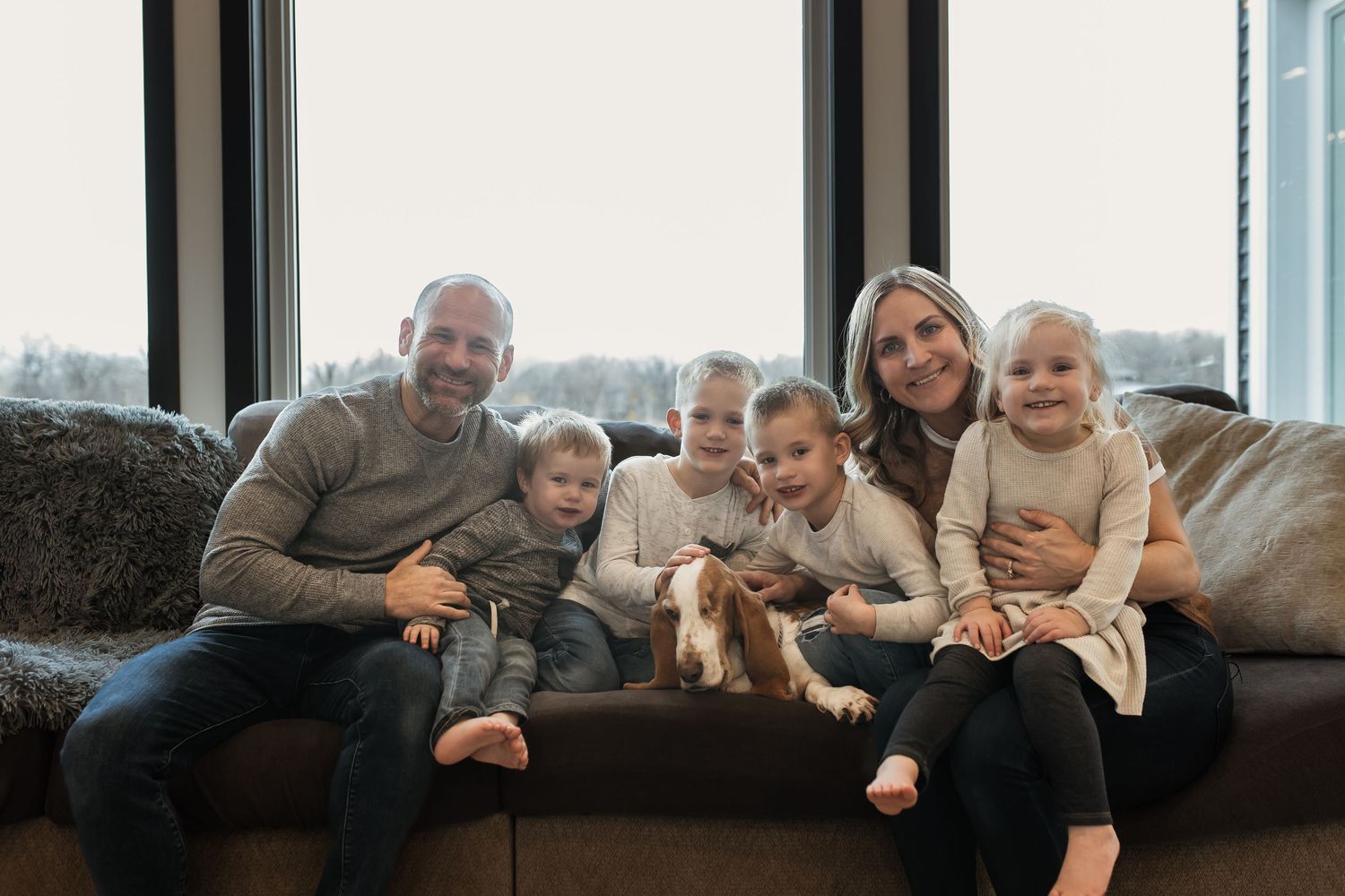 Cheerful family of six with a pet dog seated on a sofa by large windows in a bright living room.