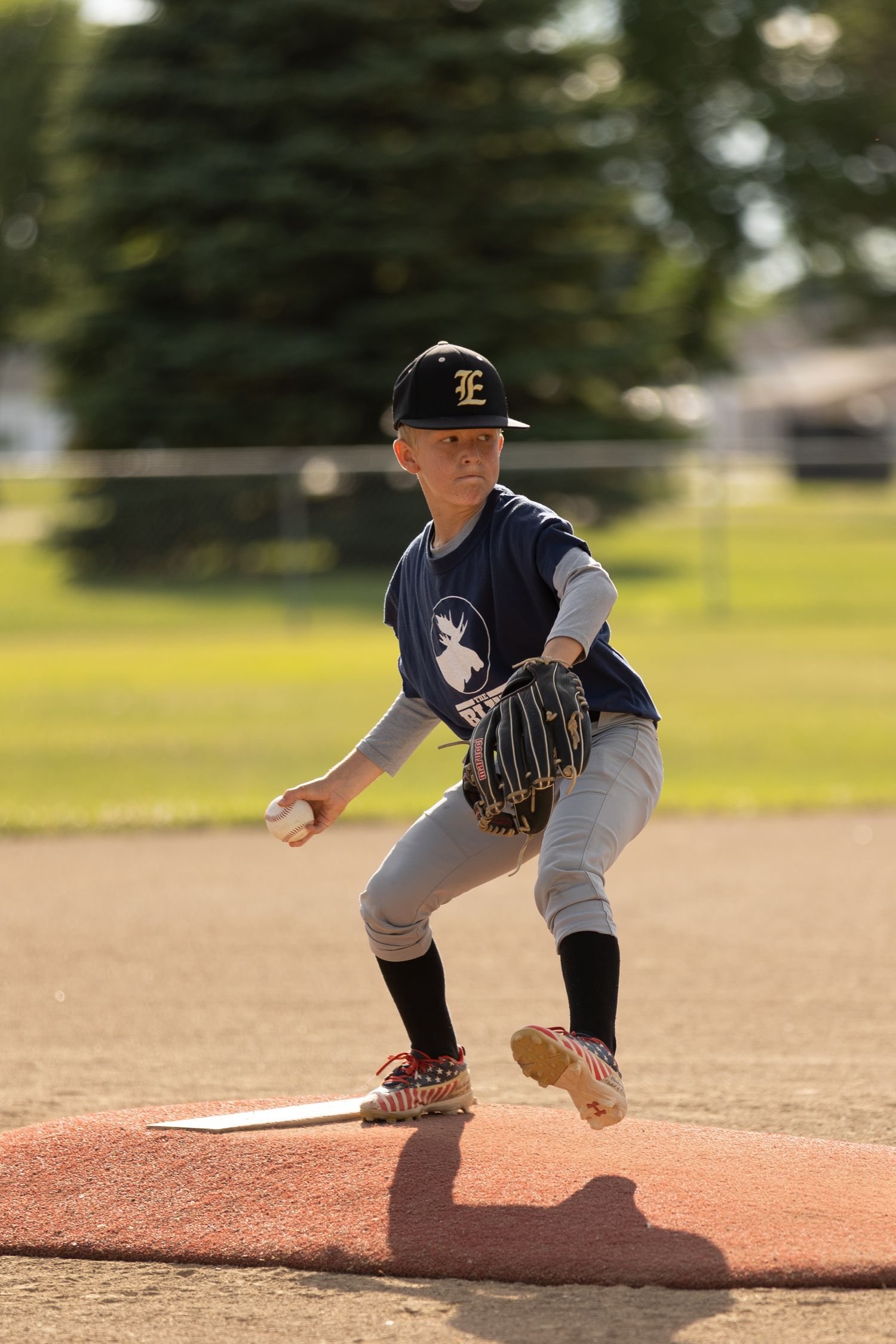 Young baseball pitcher in navy E cap winding up on pitching mound.