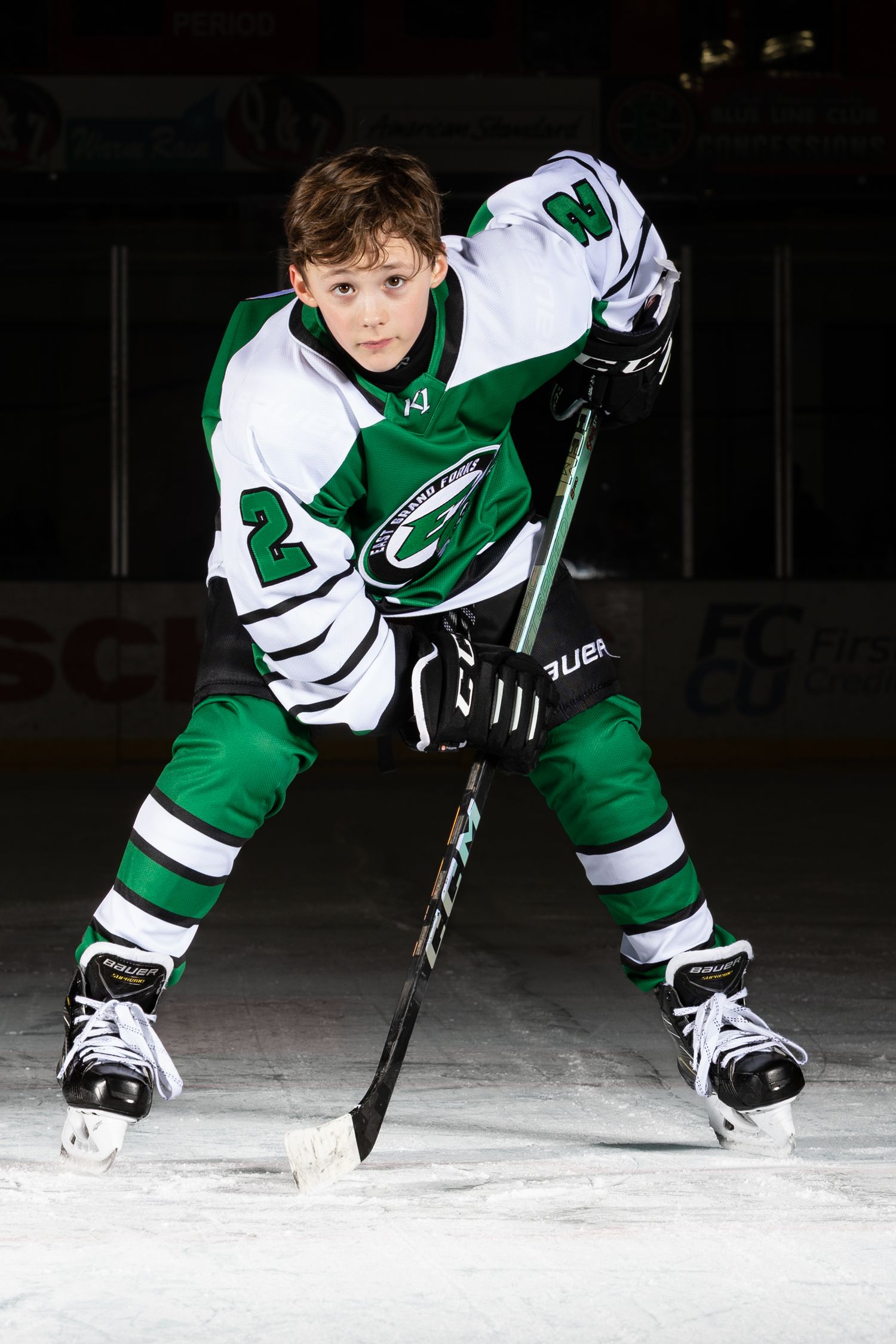 Young hockey player in green and white uniform posing with stick on ice rink.