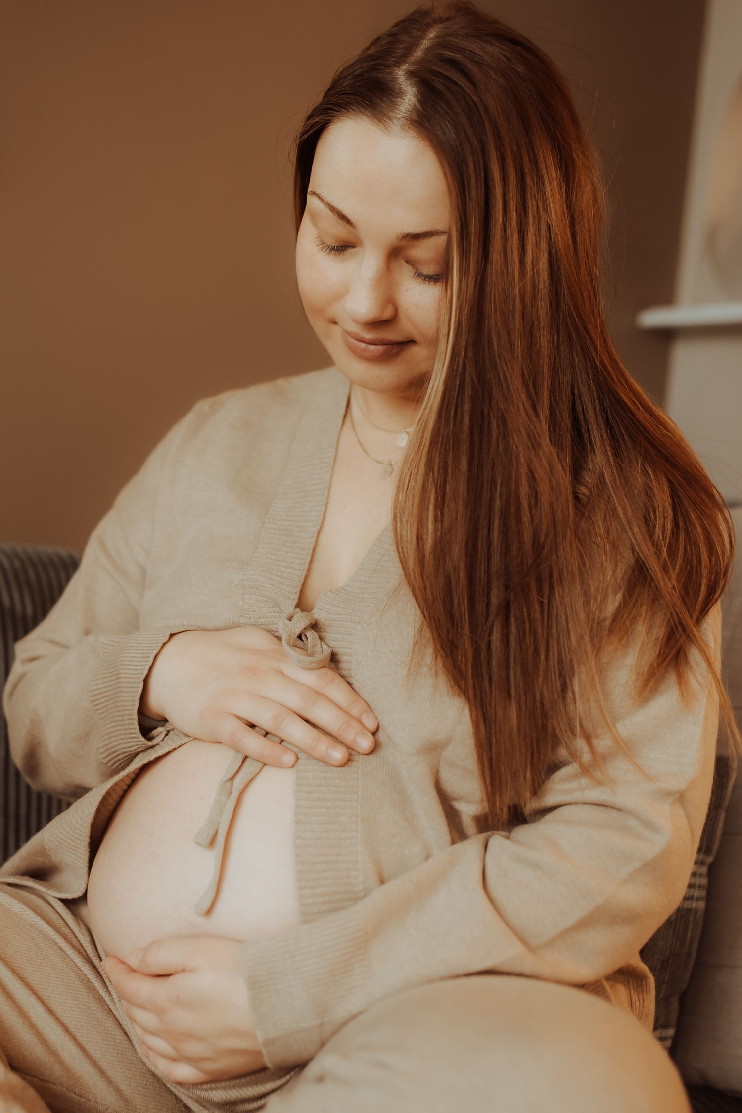 Smiling pregnant woman with long hair gently cradling her baby bump while seated indoors.