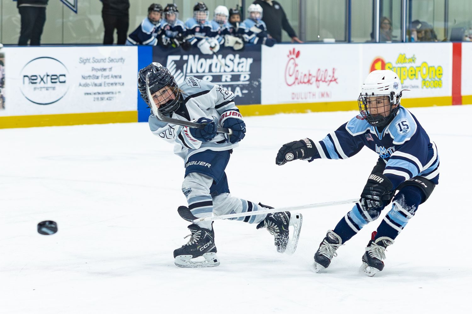 Two youth hockey players in white River uniforms battling for puck on ice.