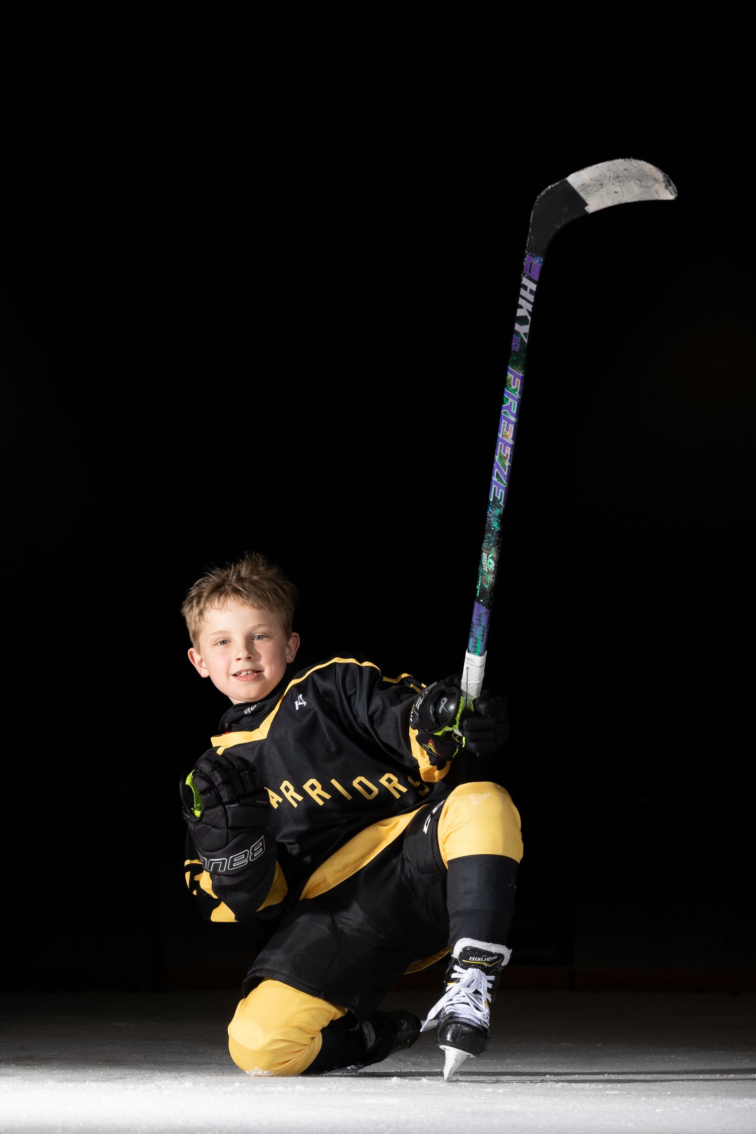 Young boy in black and gold Warrior hockey uniform kneeling on ice holding stick up.