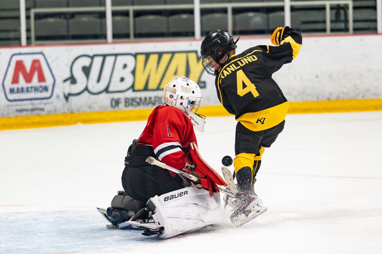 Youth hockey player attempts a shot on goal as the goalie slides to make a save in an arena game.