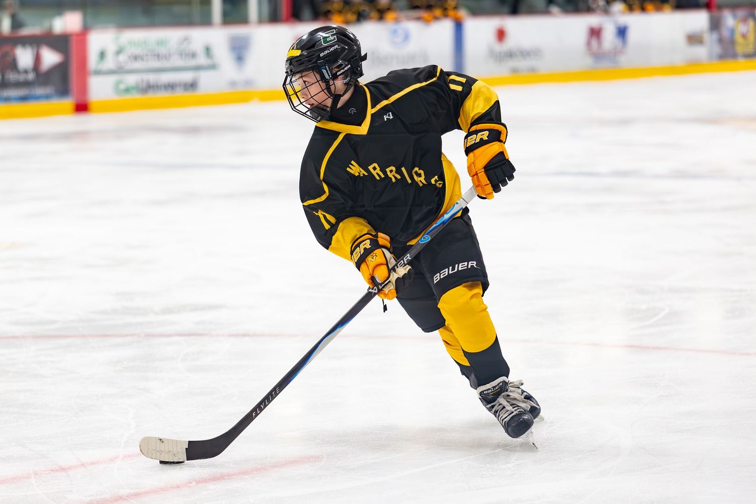 Youth hockey player in black and gold jersey skating with stick on ice rink.
