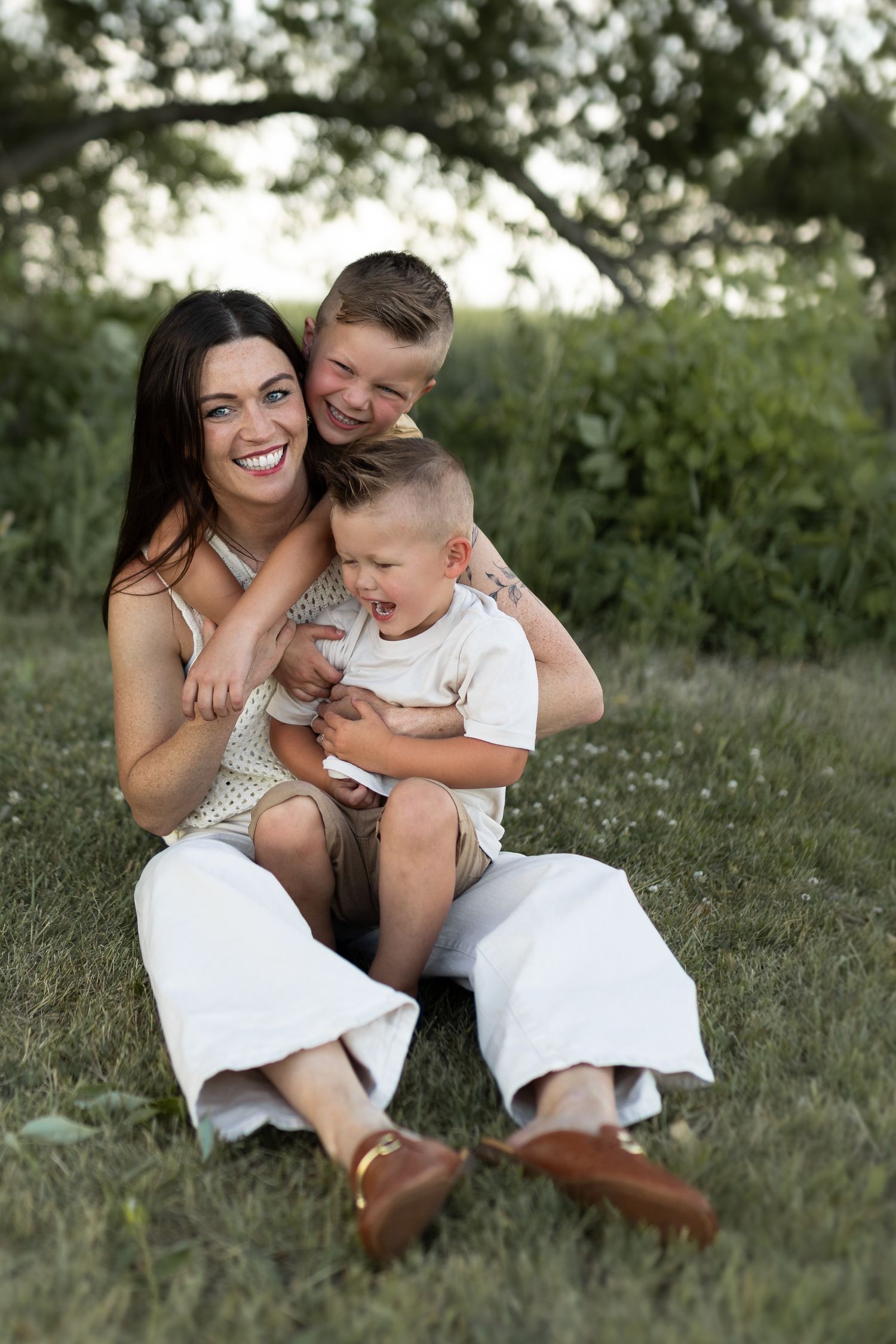 A smiling mother sits on green grass outdoors with her two young sons hugging her, enjoying a joyful family moment.