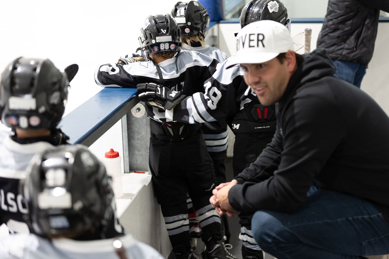Hockey coach crouching down to talk with young players in black and white uniforms on the bench during a game.