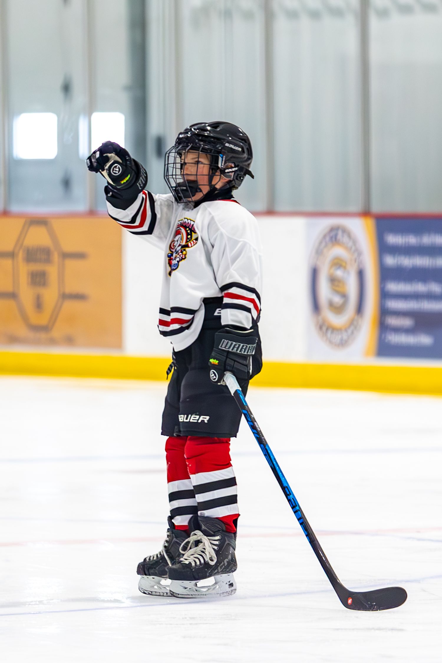 Young youth hockey player in white uniform raising hand while standing on ice rink.