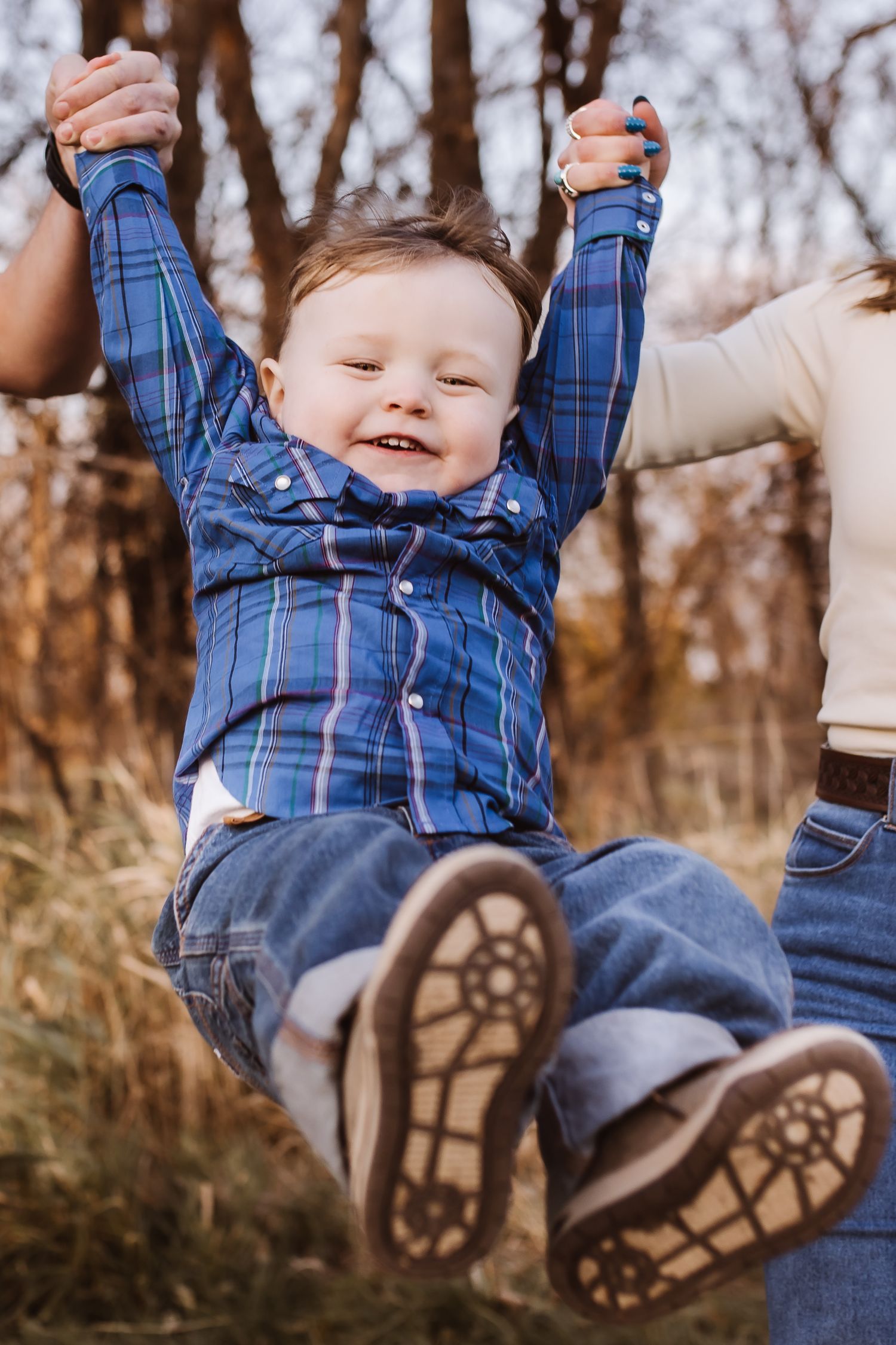 Joyful toddler boy in plaid shirt and jeans being lifted by parents outdoors in autumn setting.
