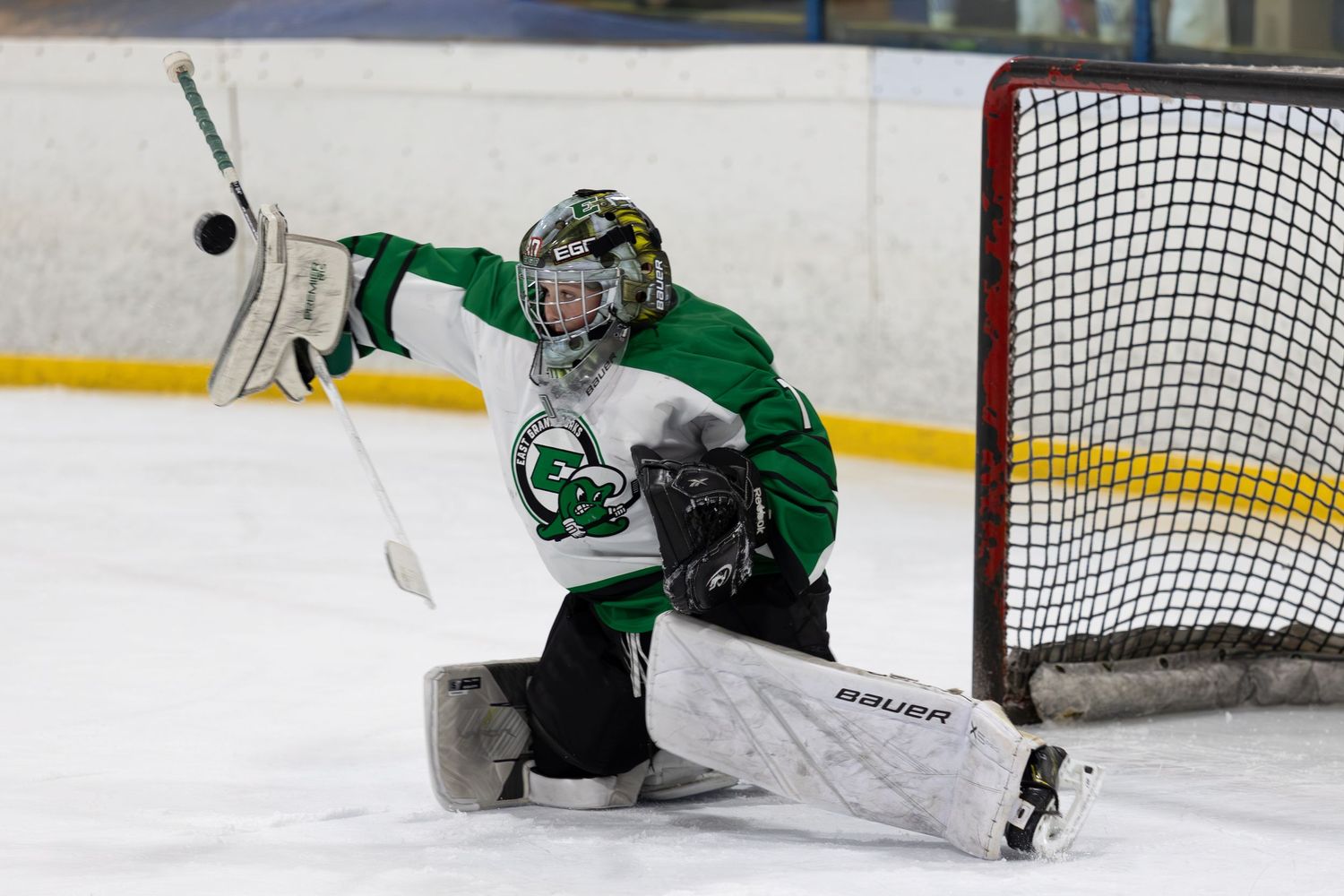 Youth hockey goalie in green uniform makes a dramatic glove save during an exciting ice hockey game.