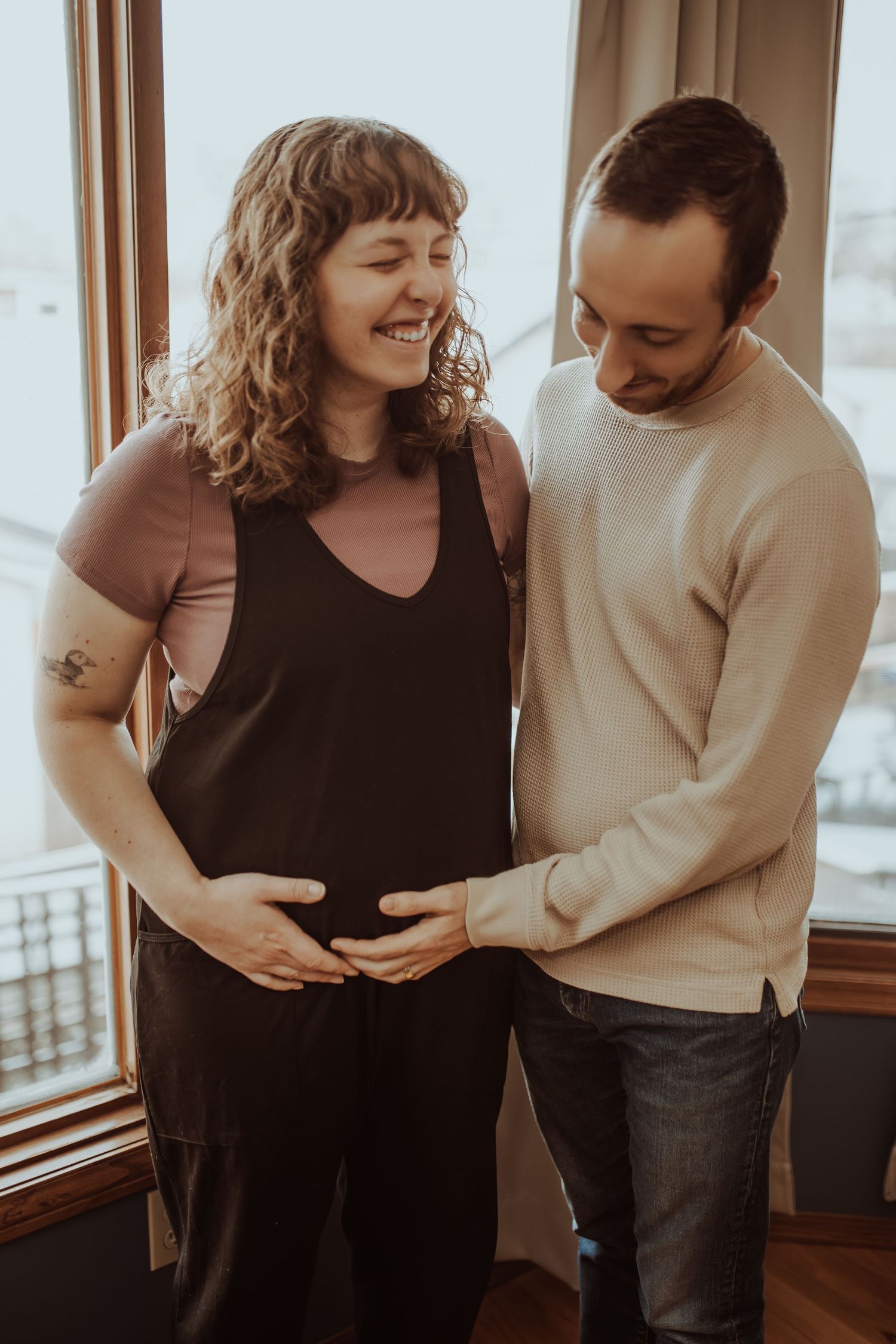 Happy expecting couple smiling and holding the pregnant woman's belly near a bright window.