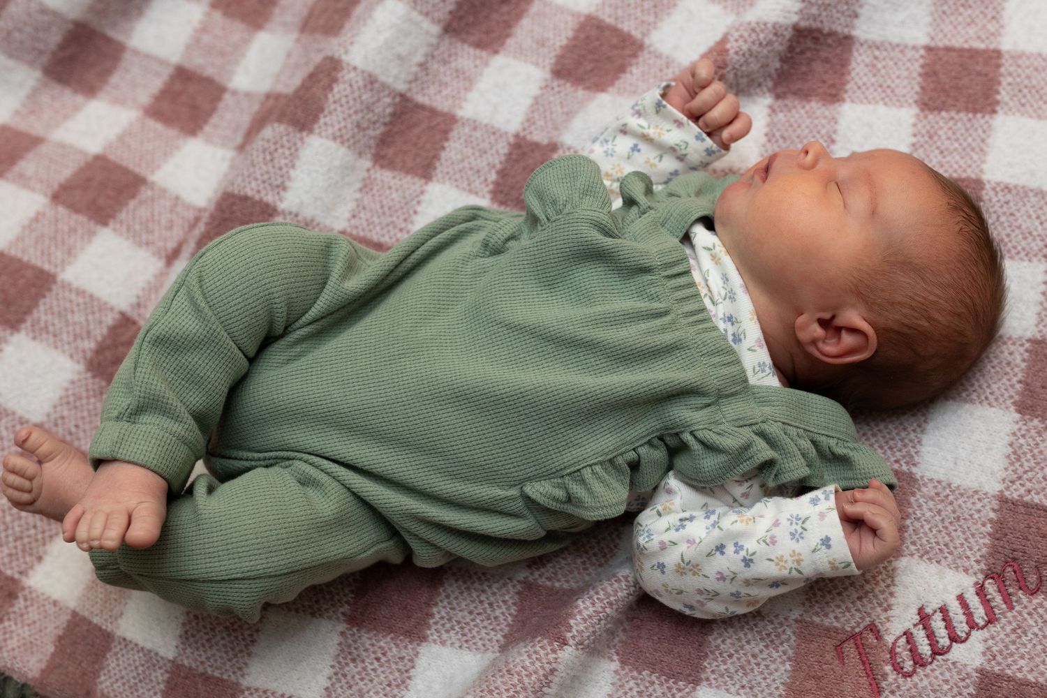 Peaceful newborn baby in a green outfit sleeping on a red plaid blanket.
