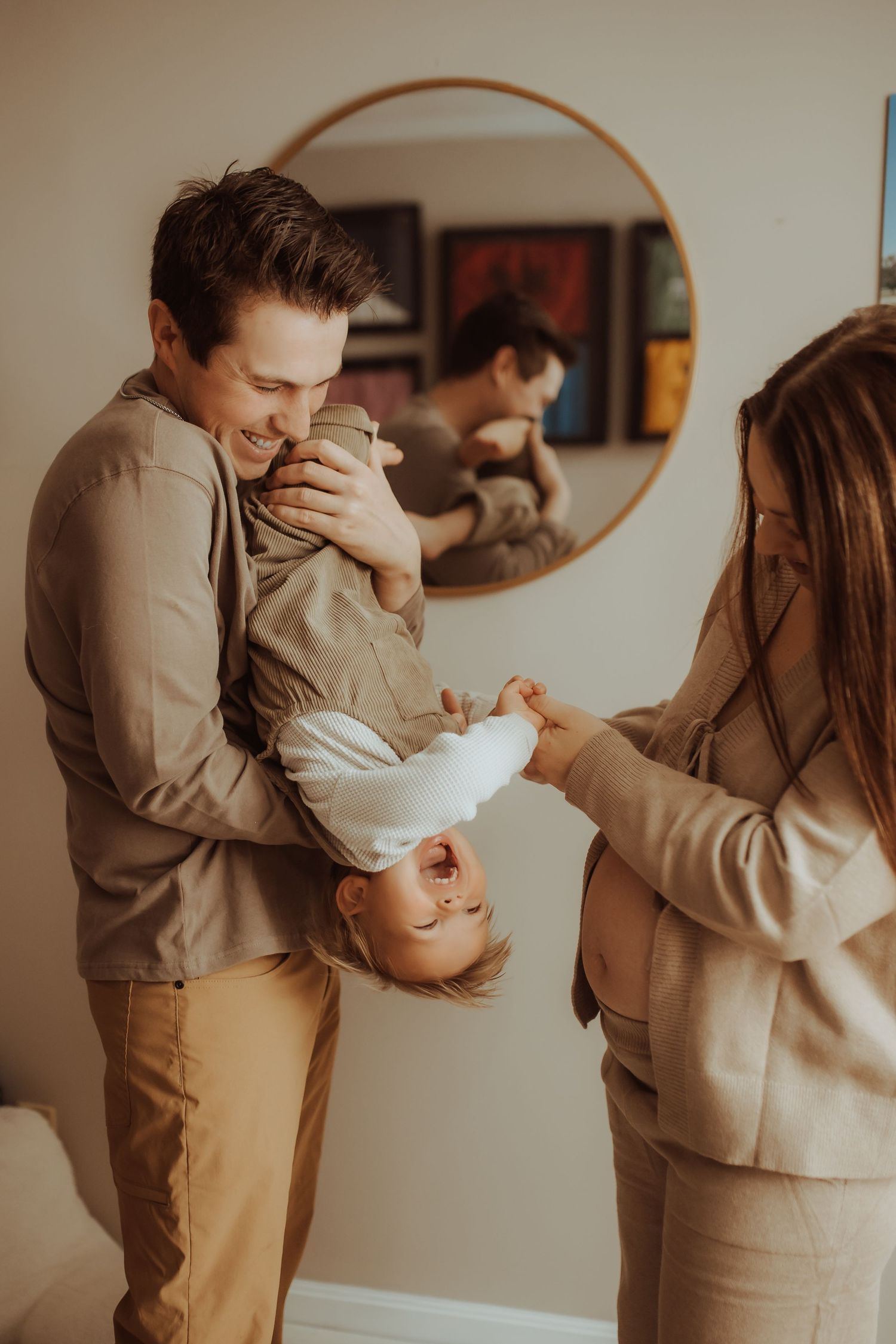 A happy family plays together at home, father holding child upside down while mother laughs, reflected in round mirror.