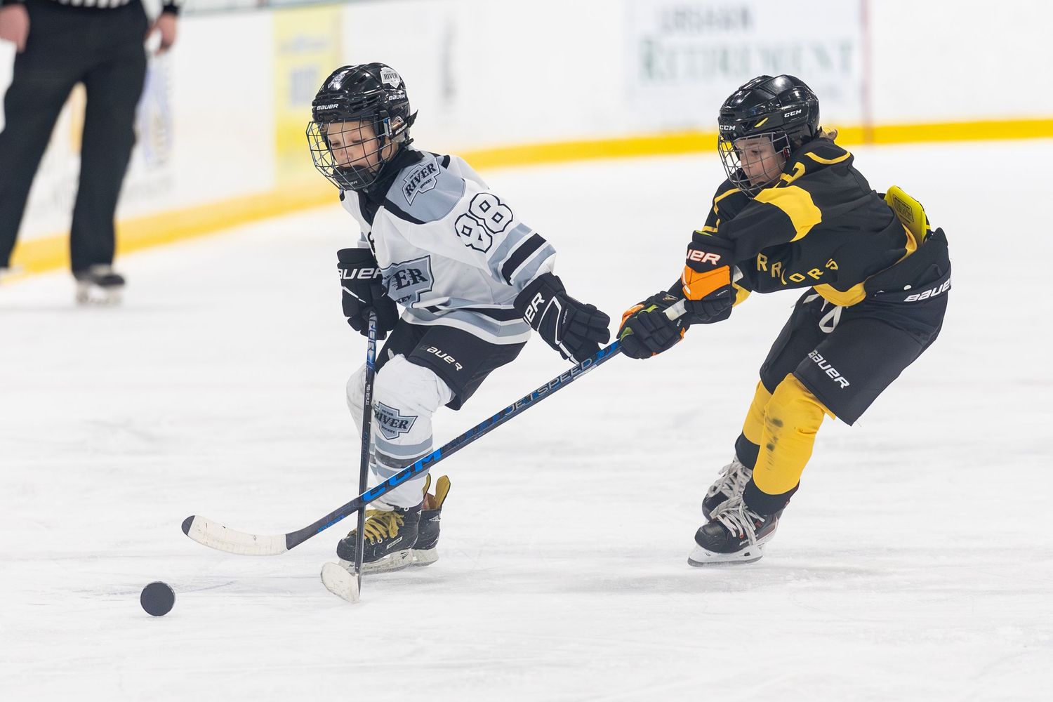 Two youth hockey players in black and white uniforms competing for puck on ice.