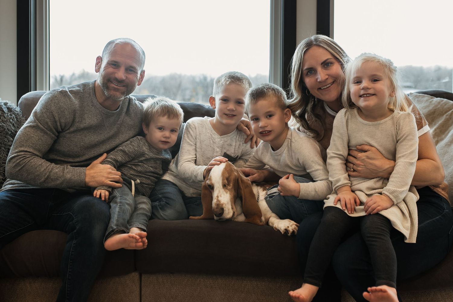 Family of six sitting on a cozy sofa with their Basset Hound dog, smiling warmly together indoors.