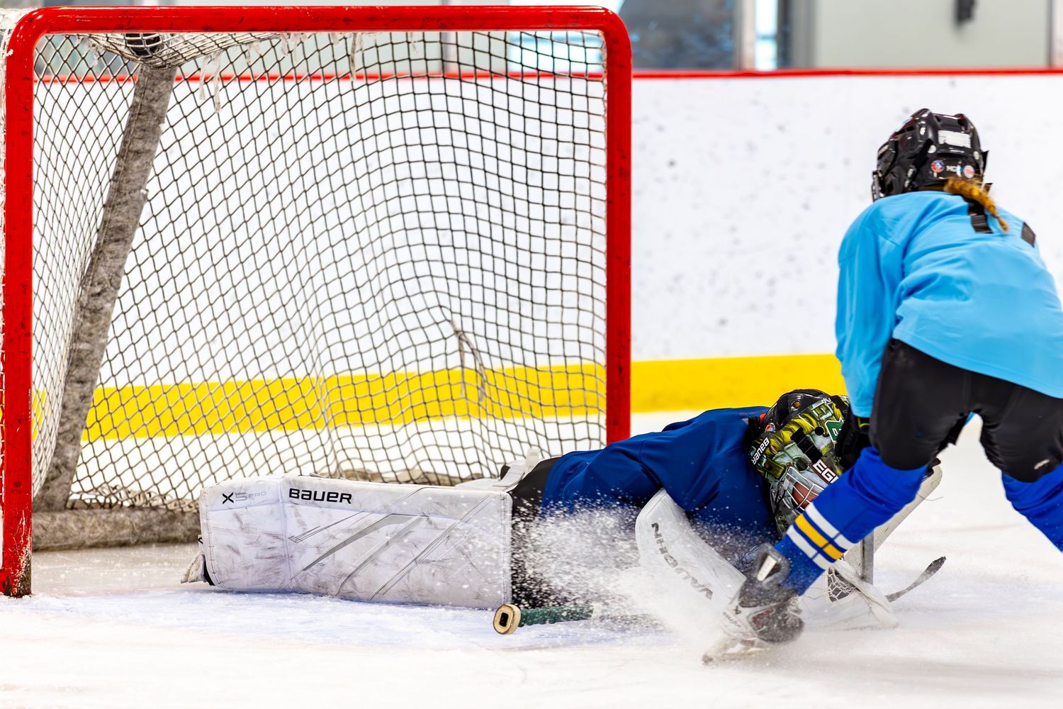 Youth hockey goalie making a save sliding on ice as player scores near the net.