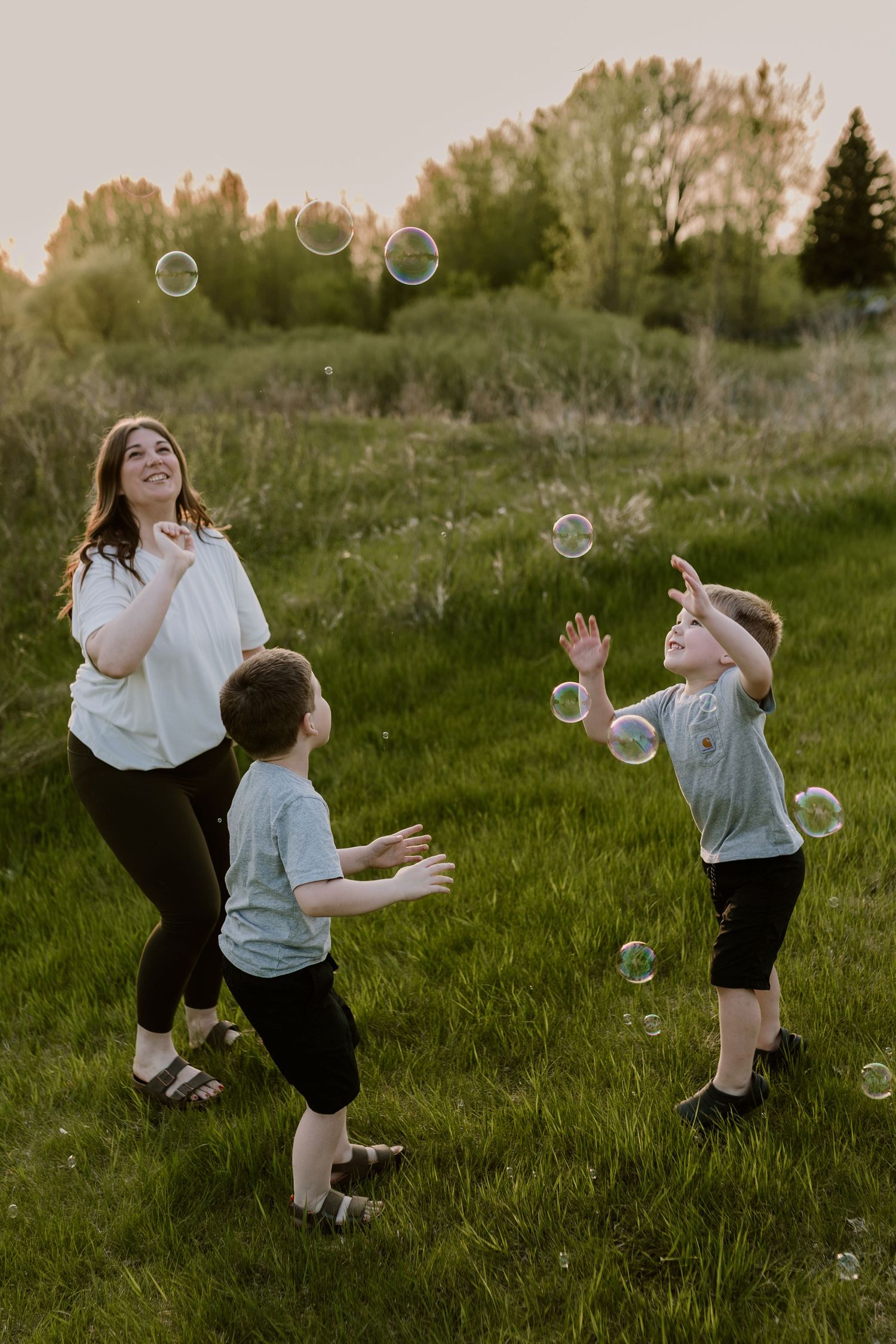 A mother and two young boys playing with bubbles in a green field at golden hour sunset.