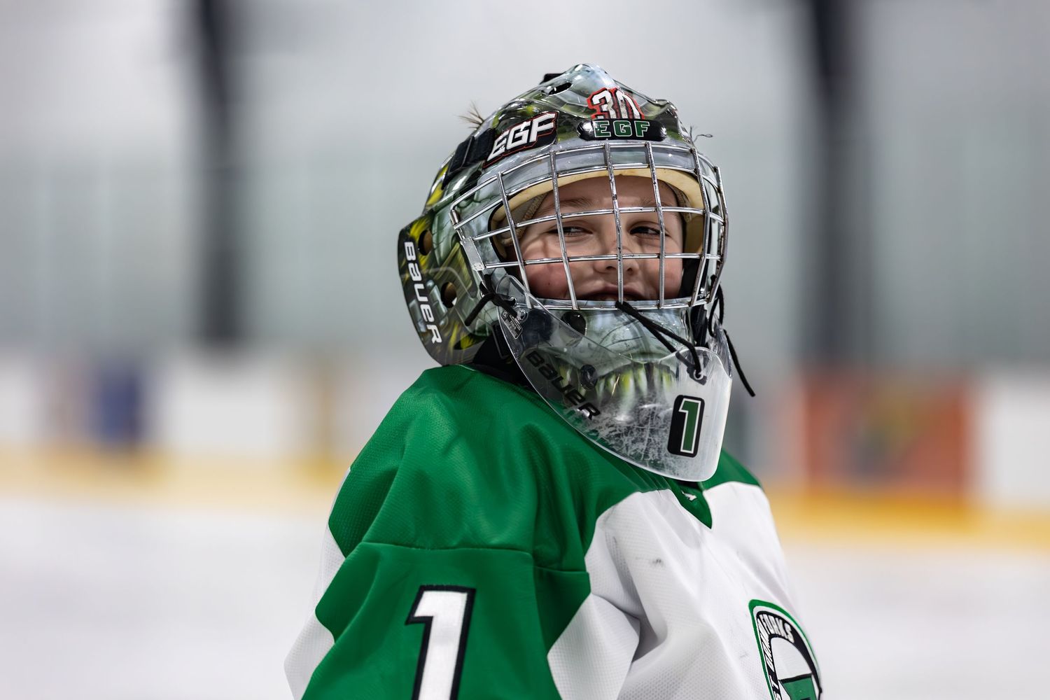 Young goalie in green hockey jersey and decorated mask smiling on ice rink.