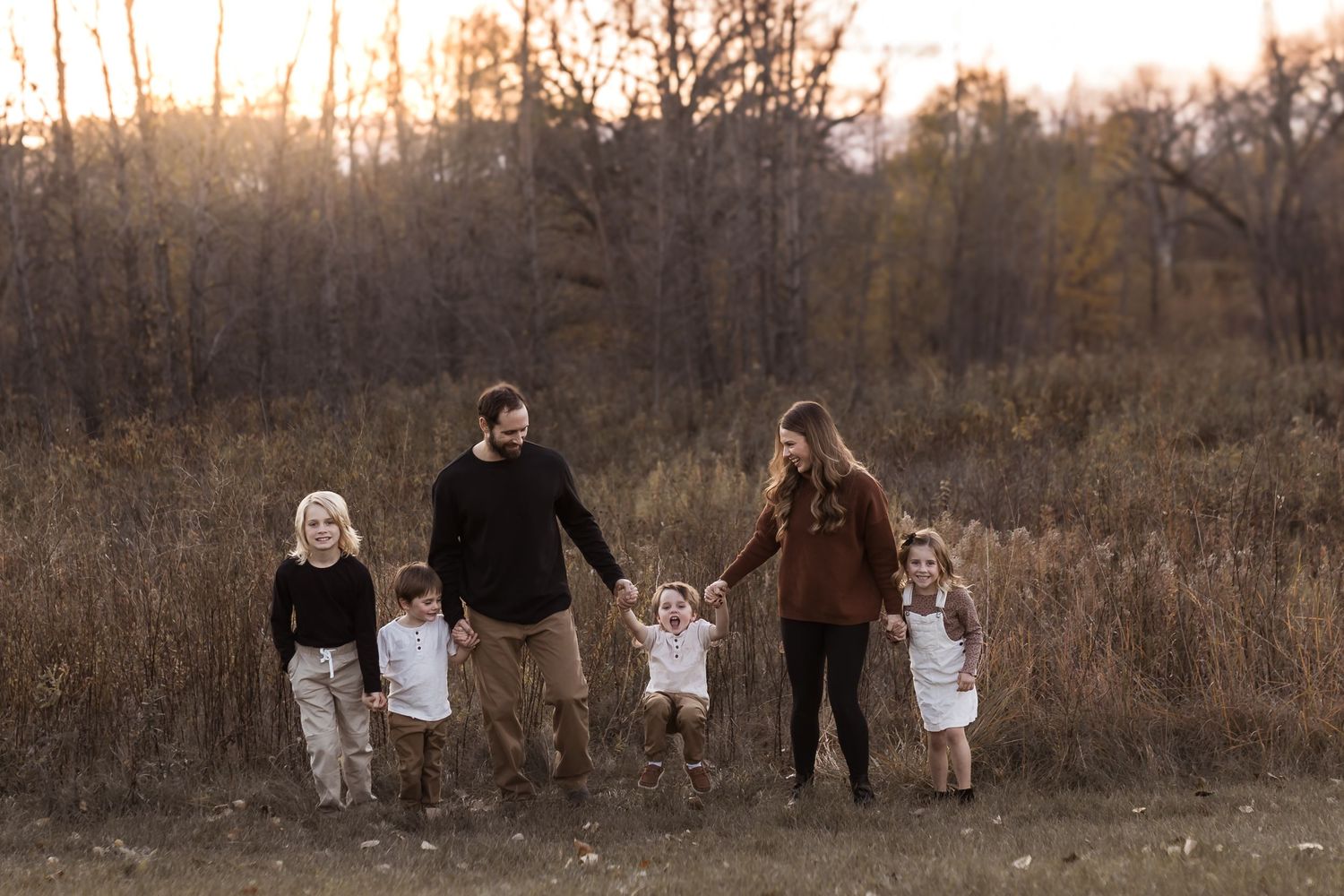 Family of five walking through golden autumn field at sunset, parents holding hands with three young children.