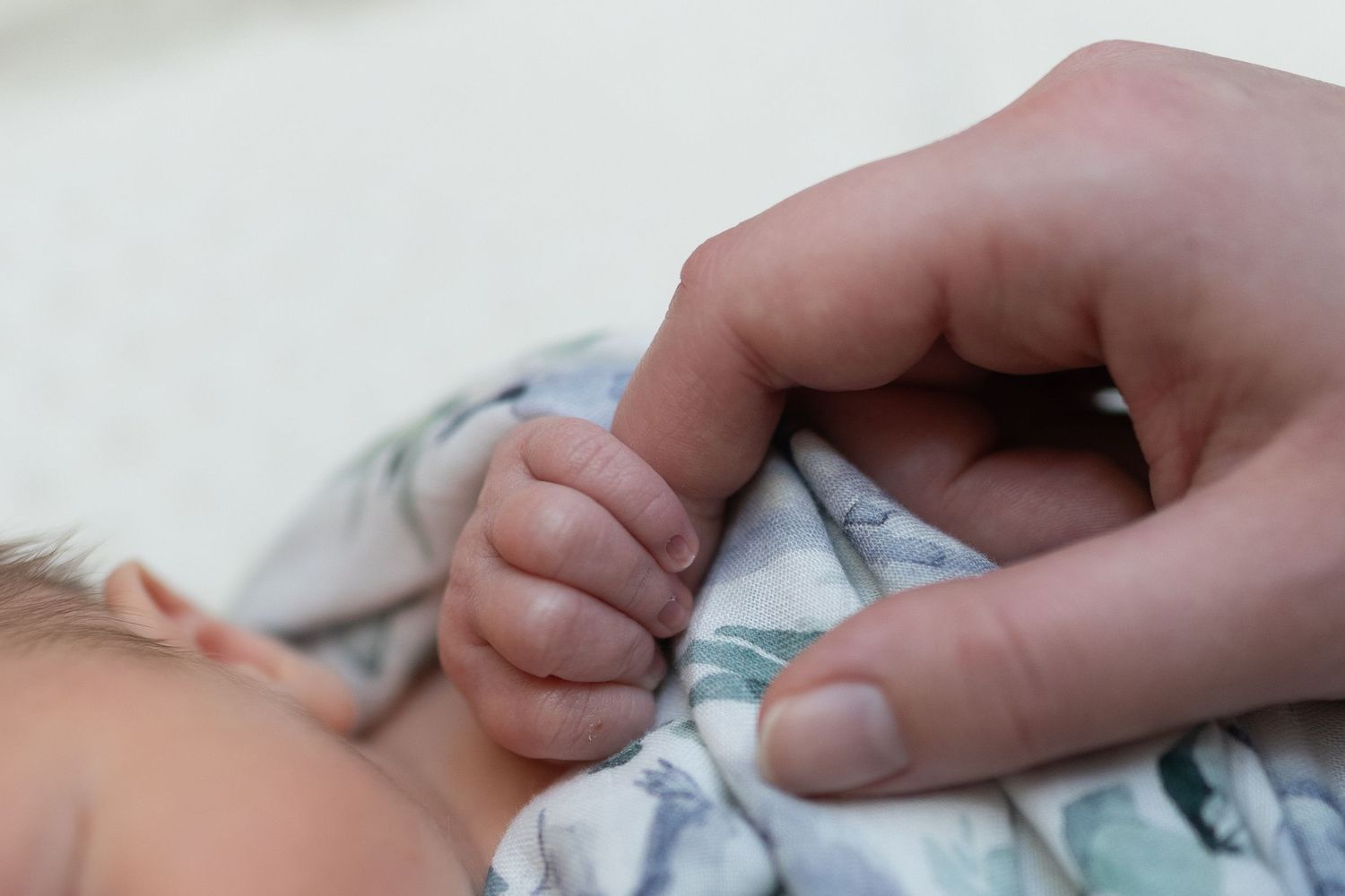 Adult finger gently held by a sleeping newborn baby's tiny hand on a floral blanket.