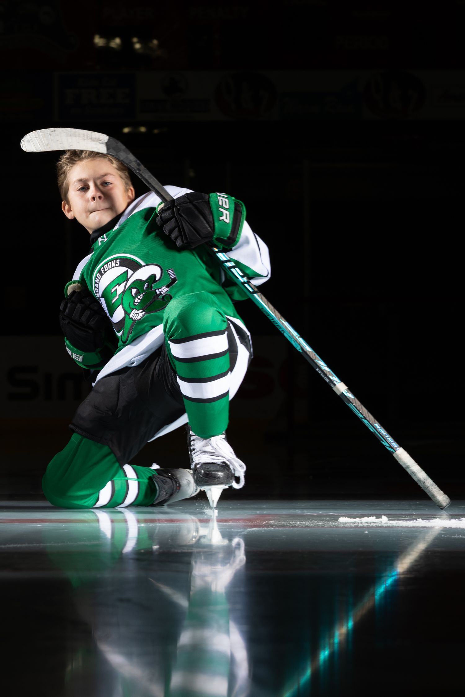 Young hockey player in green uniform kneeling dramatically on ice rink in dark arena.