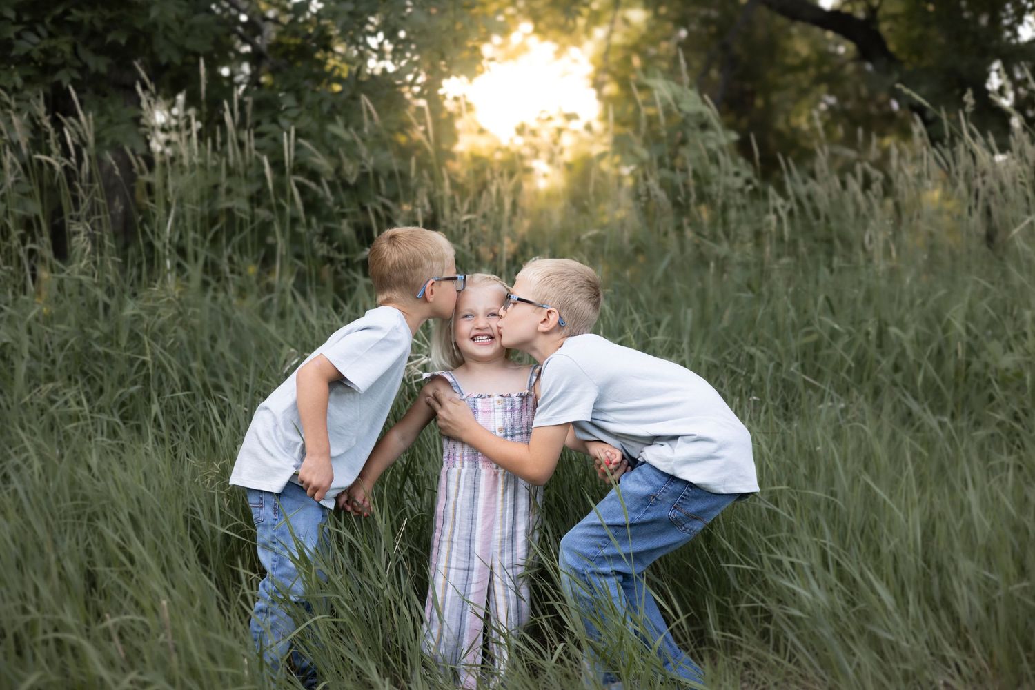 Two boys kissing a smiling girl on the cheeks in a sunlit green field at golden hour.