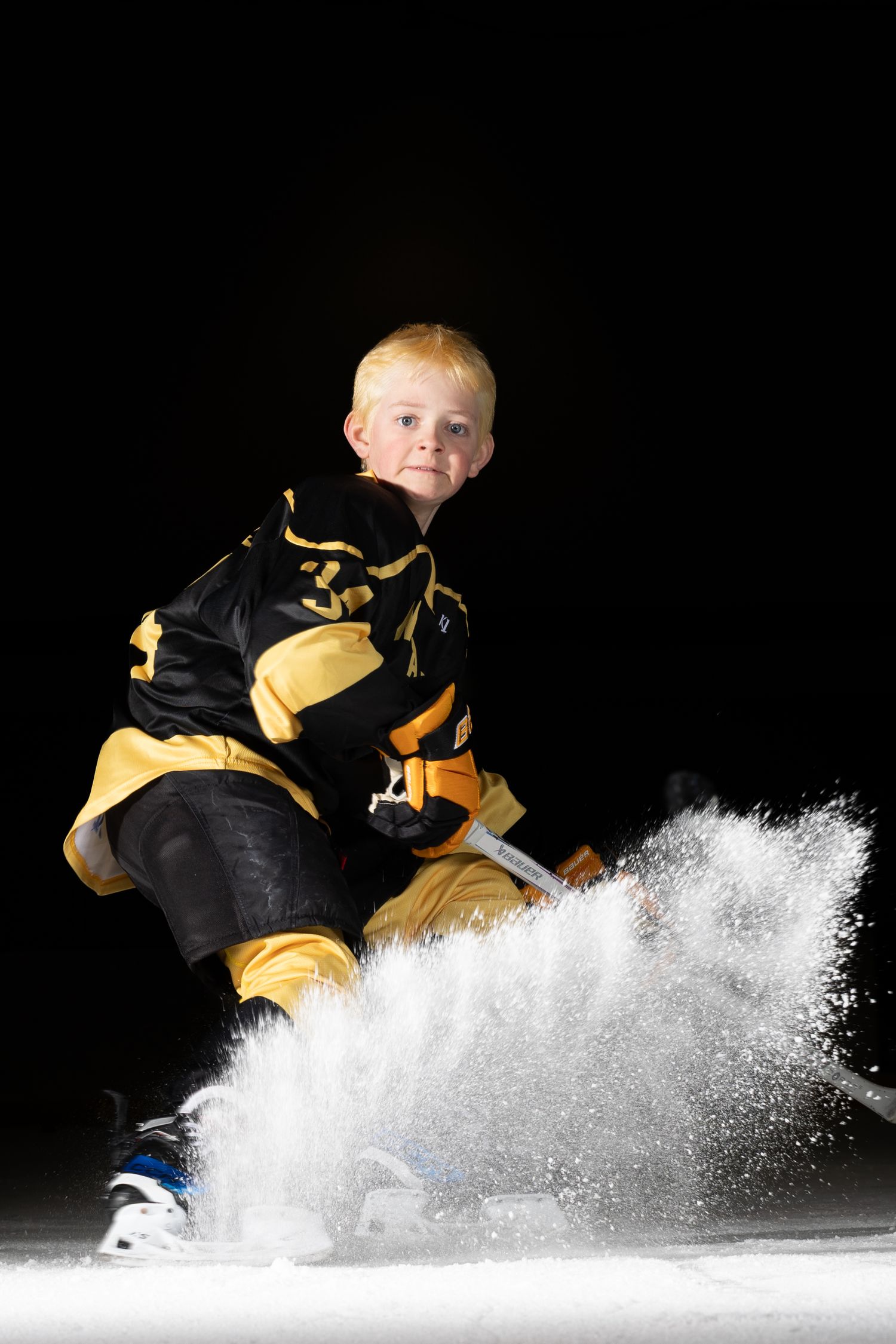 Young hockey player in black and gold uniform spraying ice while stopping on skates.