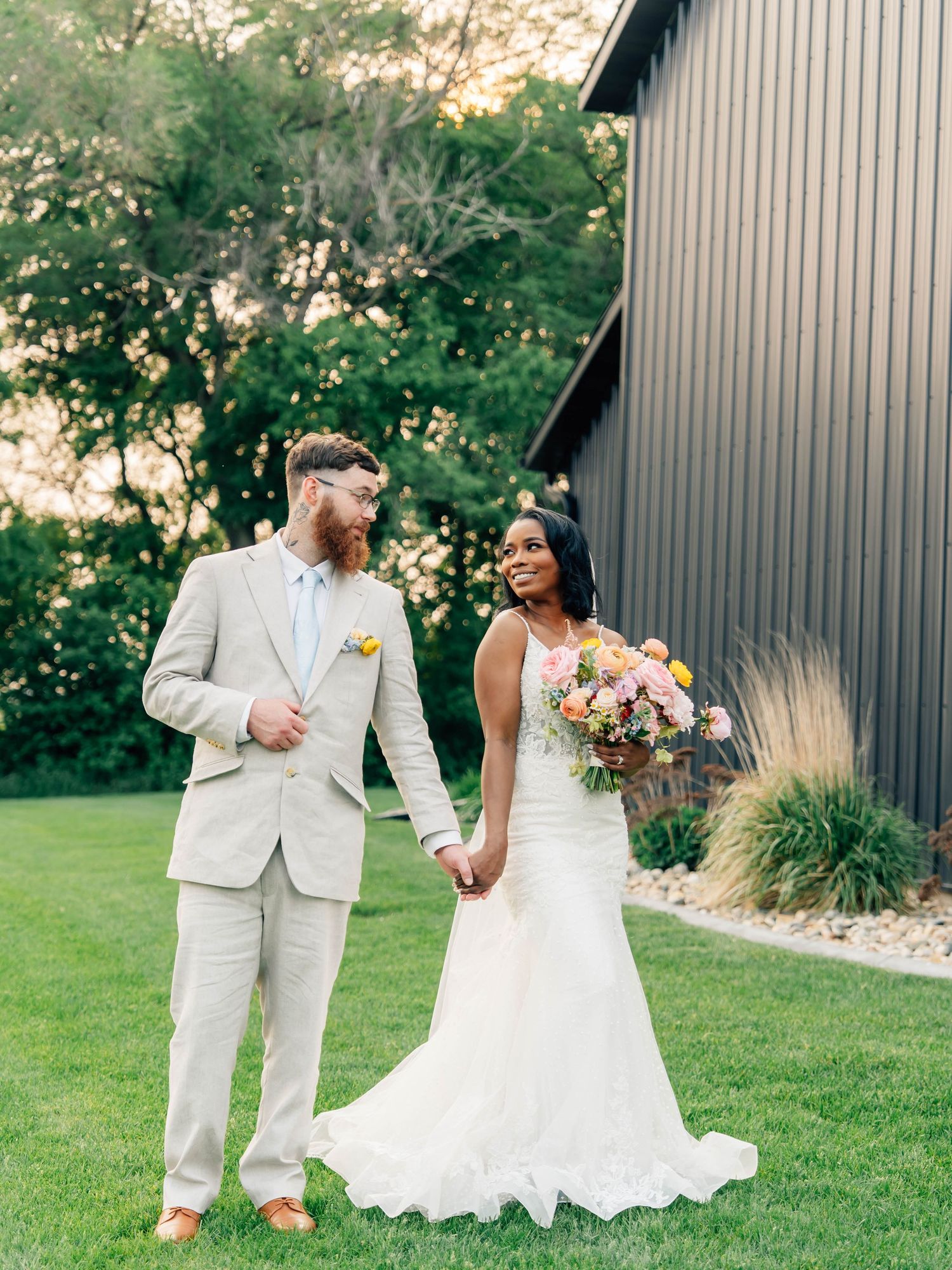 Elegant garden wedding portrait with couple in light suit and fitted lace gown holding colorful bouquet.