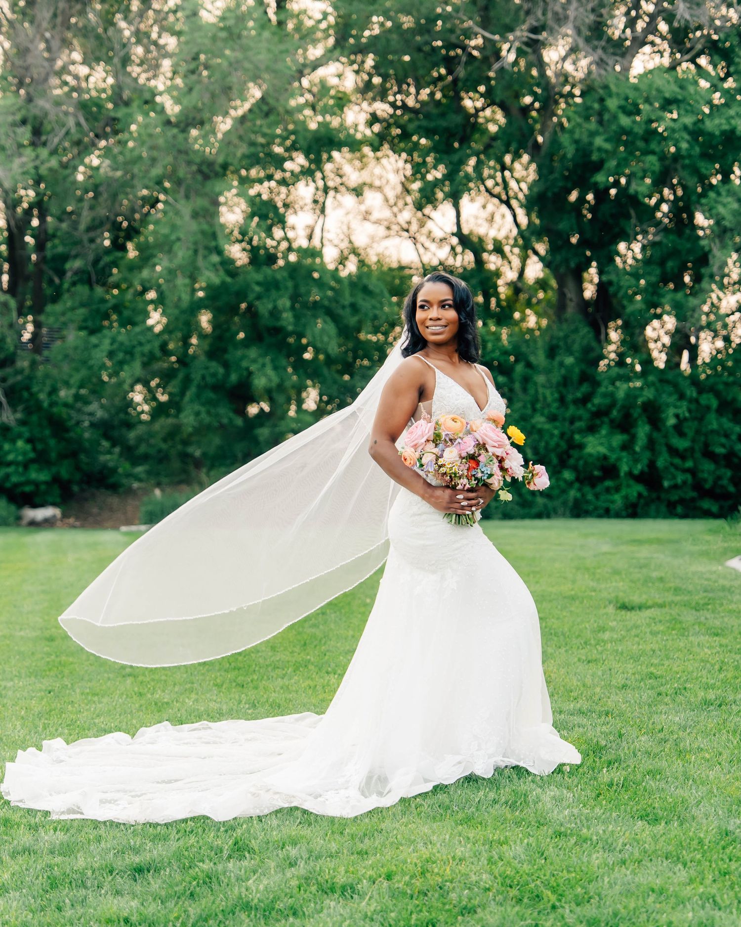 Elegant bride twirls in flowing white wedding gown against lush green outdoor backdrop.